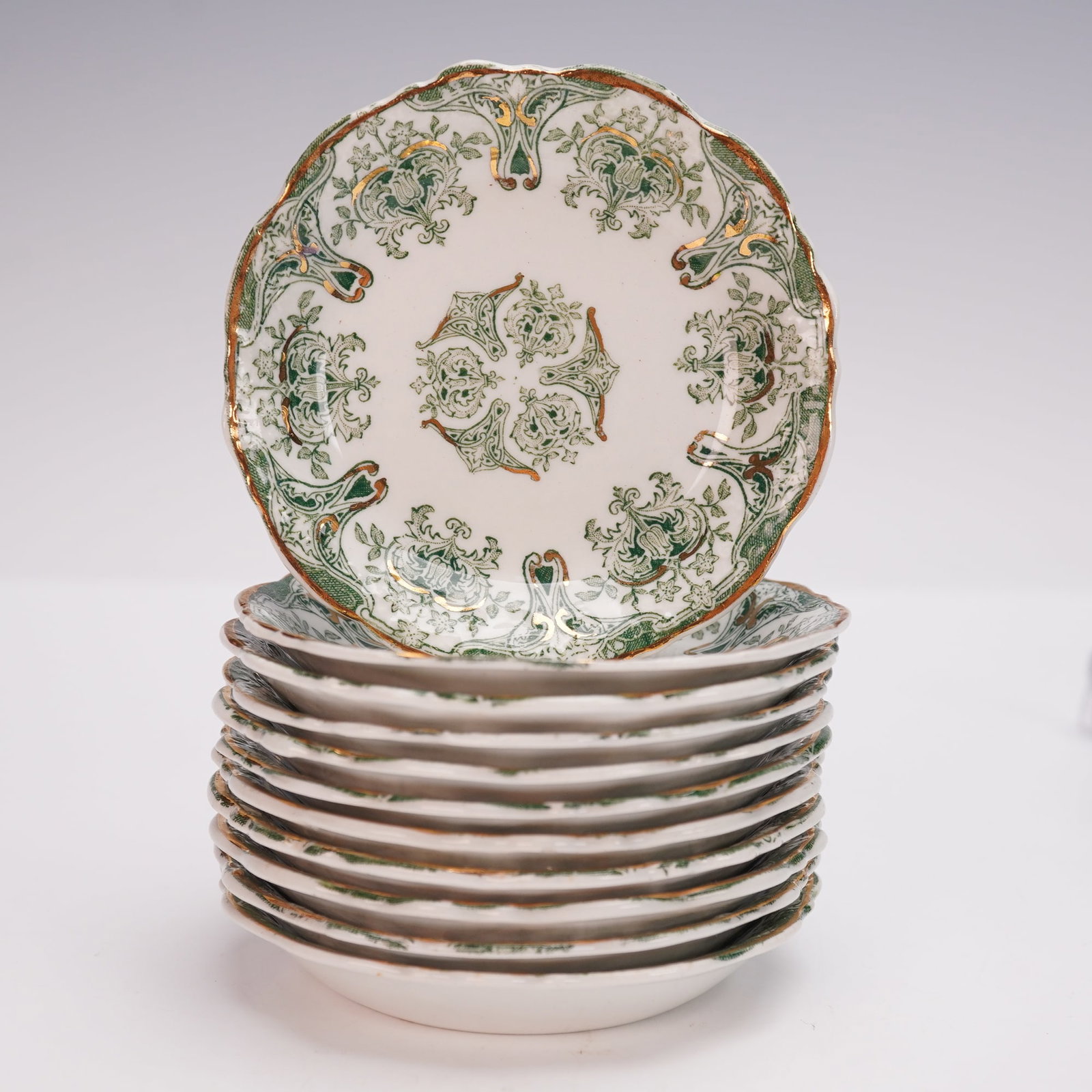 11pc J & G Meakin Hanely Genoa Flat Demitasse Saucers: Each saucer features a scalloped edge with ornate green transfer decoration accented with gilt highlights. The pattern displays intricate floral and foliate motifs with a repeating central medallion, 