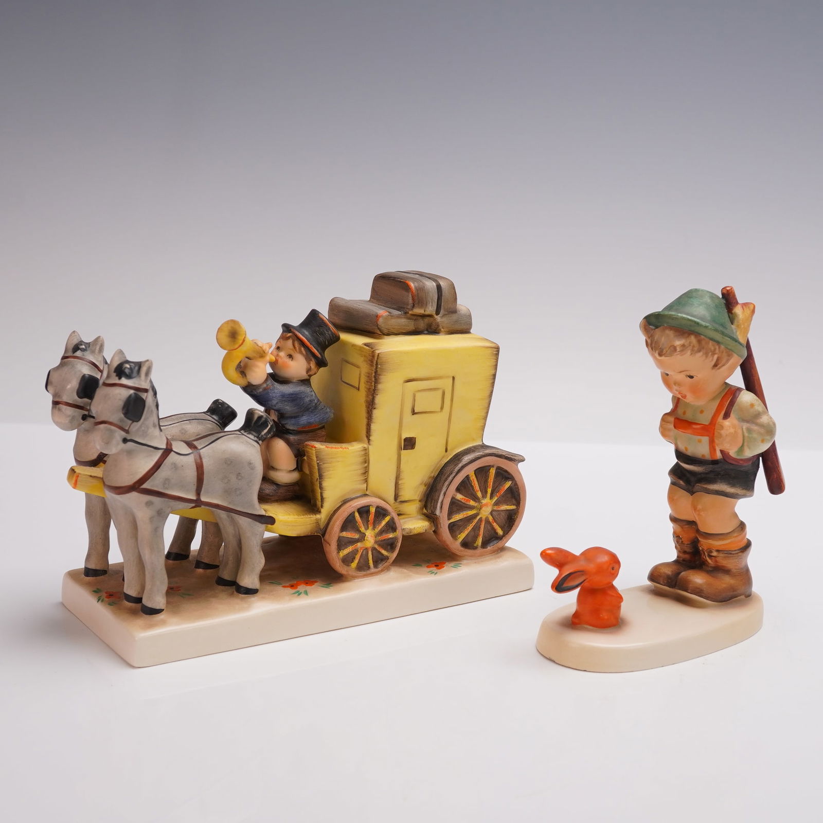 2pc Goebel Hummel Figurines, Sensitive Hunter and Mail Is Here: Two porcelain figurines including Sensitive Hunter (6/0) featuring a young boy with a walking stick and rabbit companion, and Mail Is Here (266) depicting a coachman with trumpet driving a yellow stag