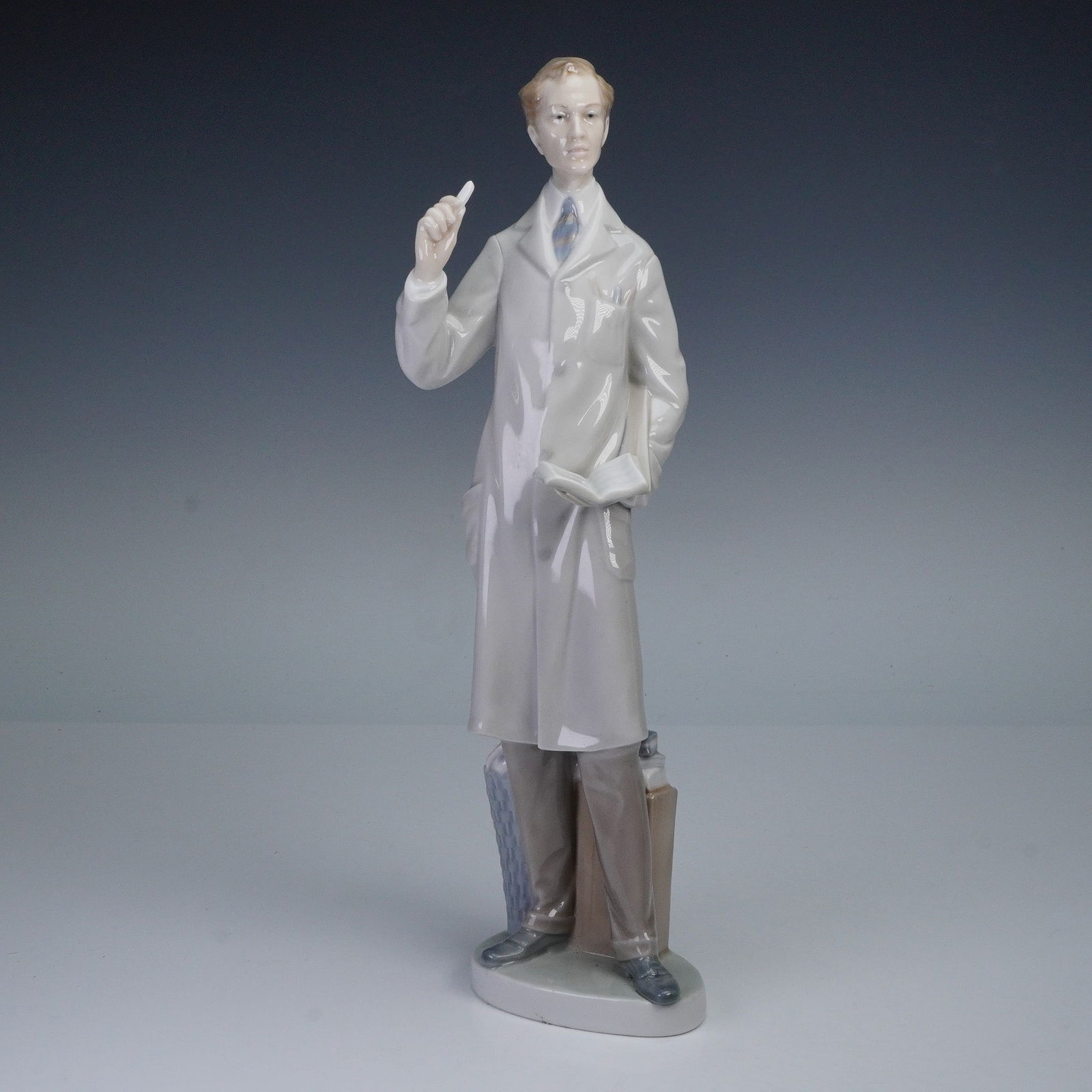 Lladro Porcelain Figurine, The Teacher 1004801: A Lladro porcelain figurine titled The Teacher, model number 1004801, hand made in Spain. The piece depicts a male teacher wearing a long white coat, holding a piece of chalk in one hand with the othe