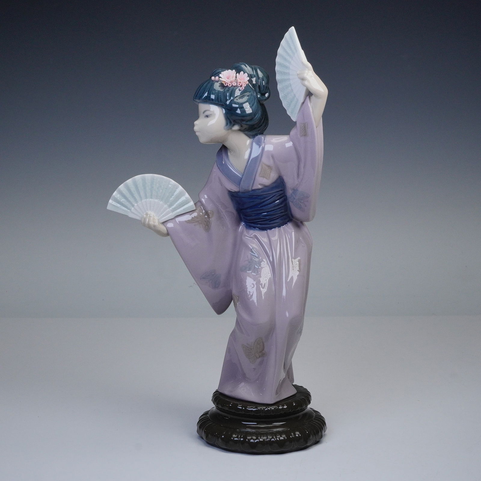Lladro Porcelain Figurine, Madame Butterfly 1004991: Lladro porcelain figurine titled Madame Butterfly, executed in a glaze finish, depicting a young woman in a lilac kimono. Elegantly poised for traditional dance, she holds two open fans and pink bloss