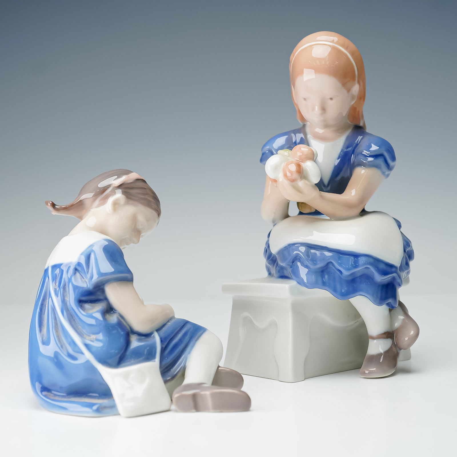 2pc Bing & Grondahl Porcelain Young Girl Figurines: A two piece grouping of Bing & Grondahl porcelain figurines including Girl Sitting with Flowers 2298 and Little Girl with Doll 1526. Each hand painted in shades of blue and white with naturalistic det