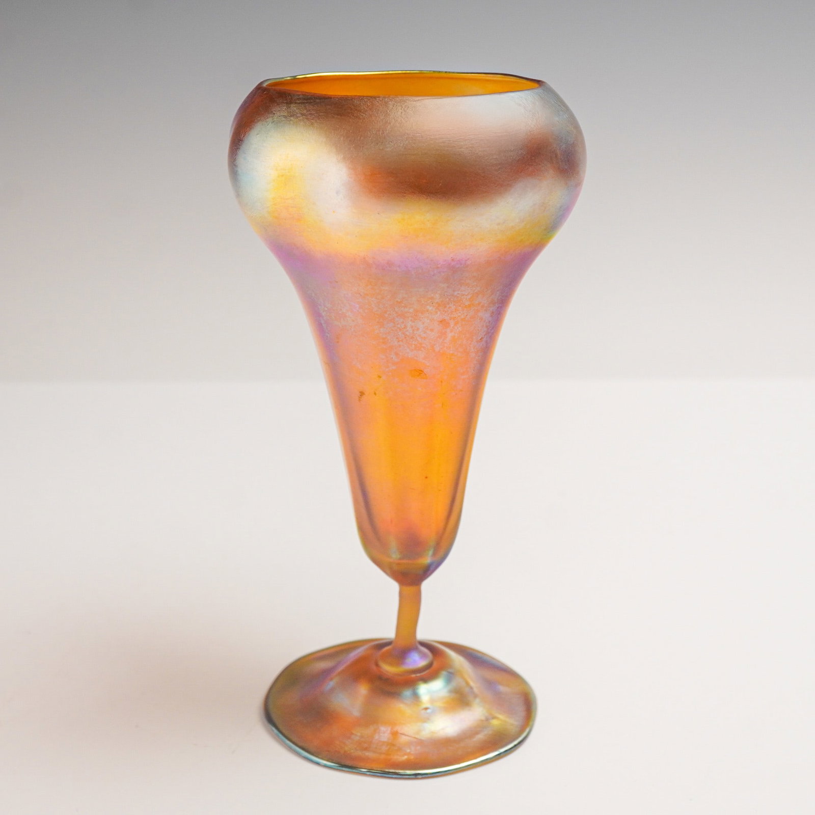 Louis Comfort Tiffany Favrile Glass Iridescent Vase: A glass vase with a bulbous rim tapering into a long slender body and resting on a broad circular base. The surface displays a luminous iridescence with golden amber, orange, and violet highlights acr