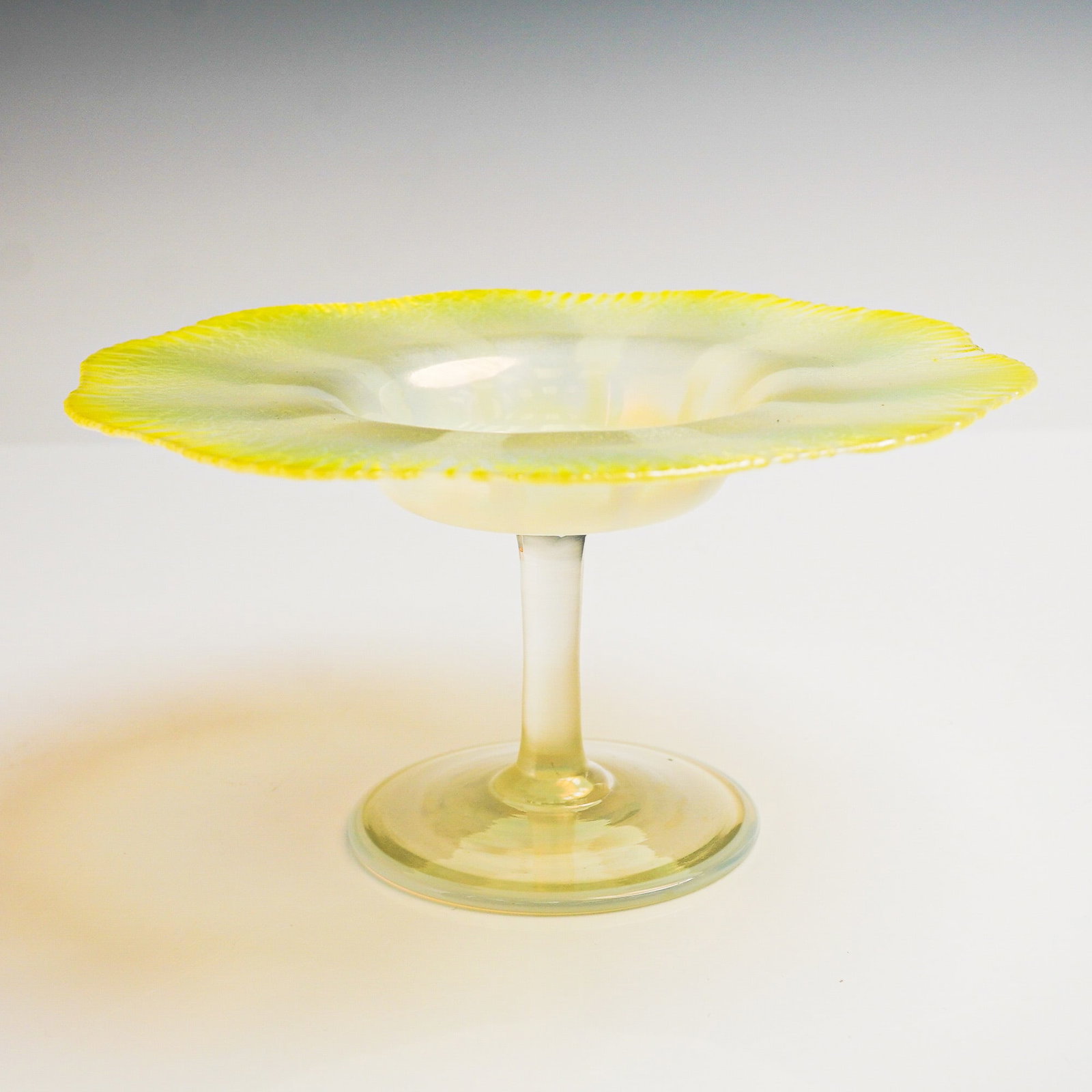 Louis Comfort Tiffany Favrile Glass Iridescent Footed Bowl: Executed in clear glass with an iridescent yellow hue along the broad ruffled rim, featuring a wide, shallow bowl raised on a slender stem and circular foot. The surface displays a subtle striated pat