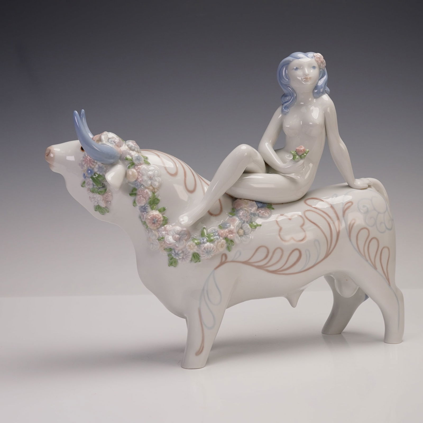 Lladro Rare Vintage Figurine, Love Europe 1001130: This sculptural porcelain piece depicts a nude female figure with flowing blue hair, seated gracefully atop a stylized bull decorated with delicate pastel floral garlands. The bull is further embellis