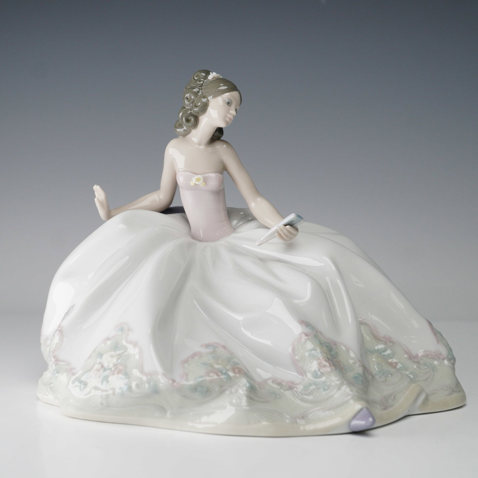 Lladro Porcelain Figurine, At the Ball 01005859: Elegant Lladro porcelain figurine titled At the Ball (01005859), designed by Jose Puche. The piece depicts a refined young woman seated gracefully with a fan in hand, her voluminous gown flowing outwa