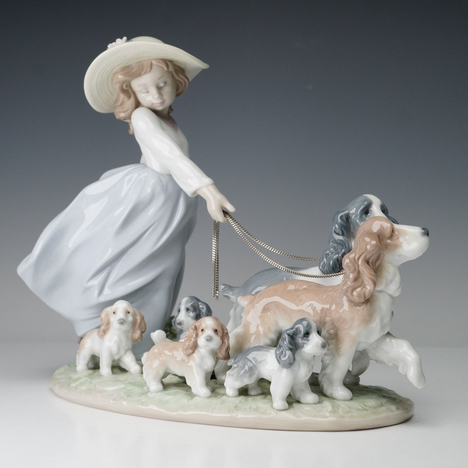 Lladro Porcelain Figurine, Puppy Parade 01006784: Charming Lladro porcelain figurine titled Puppy Parade (01006784), designed by Antonio Ramos. The piece portrays a young girl in a flowing pale blue skirt and wide-brimmed hat, gently leading a group