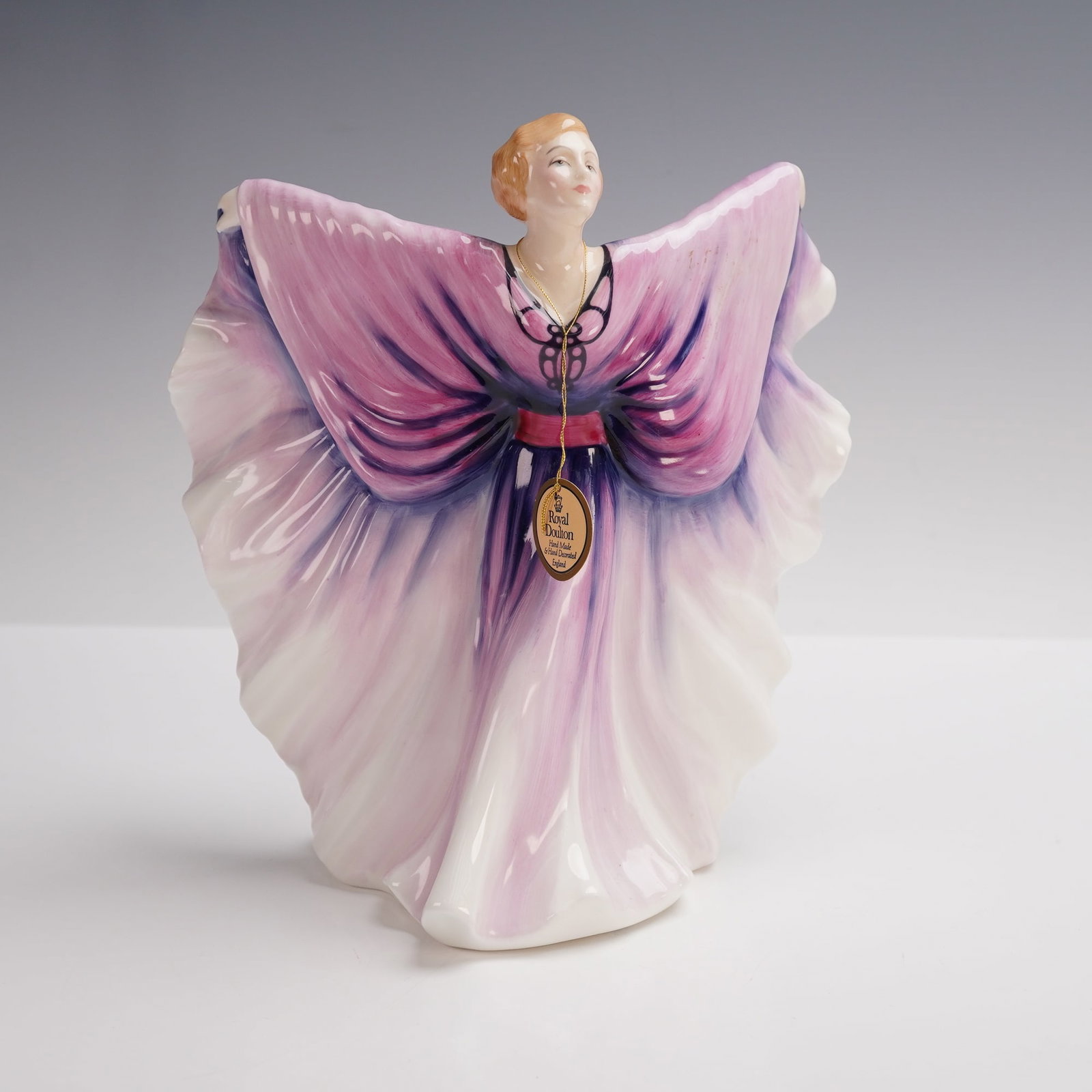 Royal Doulton Figurine, Isadora HN2938: Exuding theatrical flair, this Royal Doulton figurine is titled Isadora, model number HN2938. The design portrays a woman with outstretched arms, her gown fanned wide in flowing shades of lavender, pi