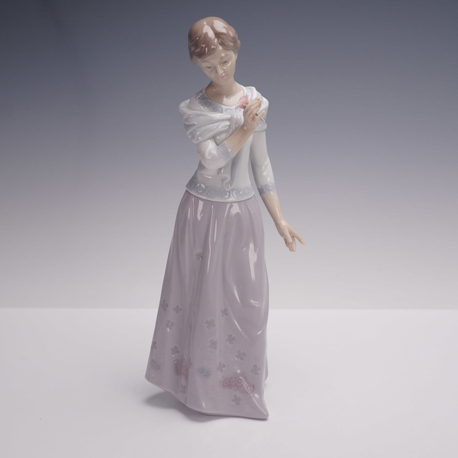 Lladro Porcelain Figurine, A Grandmother's Love 1006981: Lladro porcelain figurine titled A Grandmother's Love, model number 1006981, depicting a standing woman in a lilac floral skirt and pale blouse, holding a rose to her chest. Finished in the brand's si
