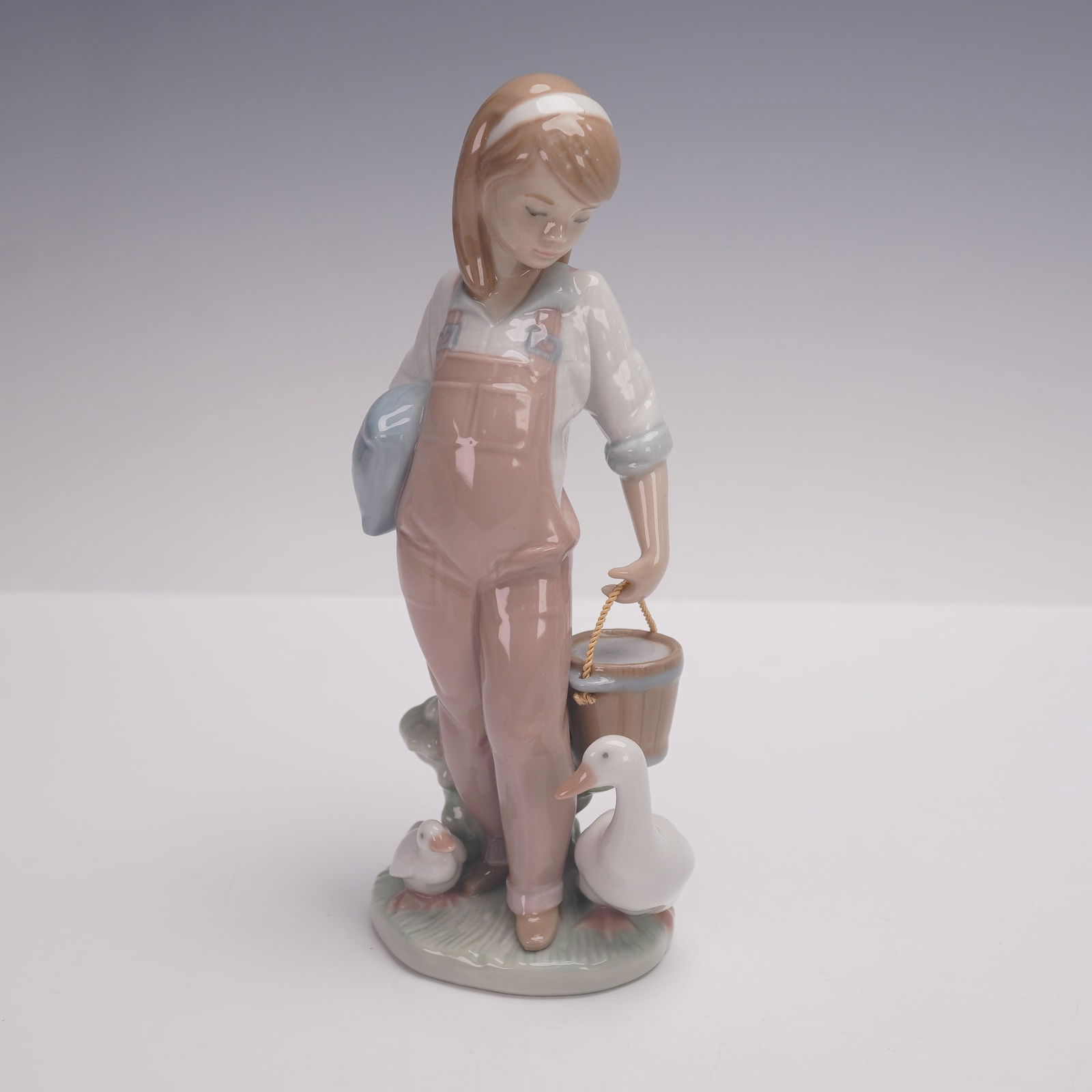 Lladro Porcelain Figurine, Saturday's Child Girl 1006022: Lladro porcelain figurine titled Saturday's Child, model number 1006022, depicting a young girl in pastel overalls holding a hat and feeding ducks beside a pail. Finished in the classic Lladro glazed