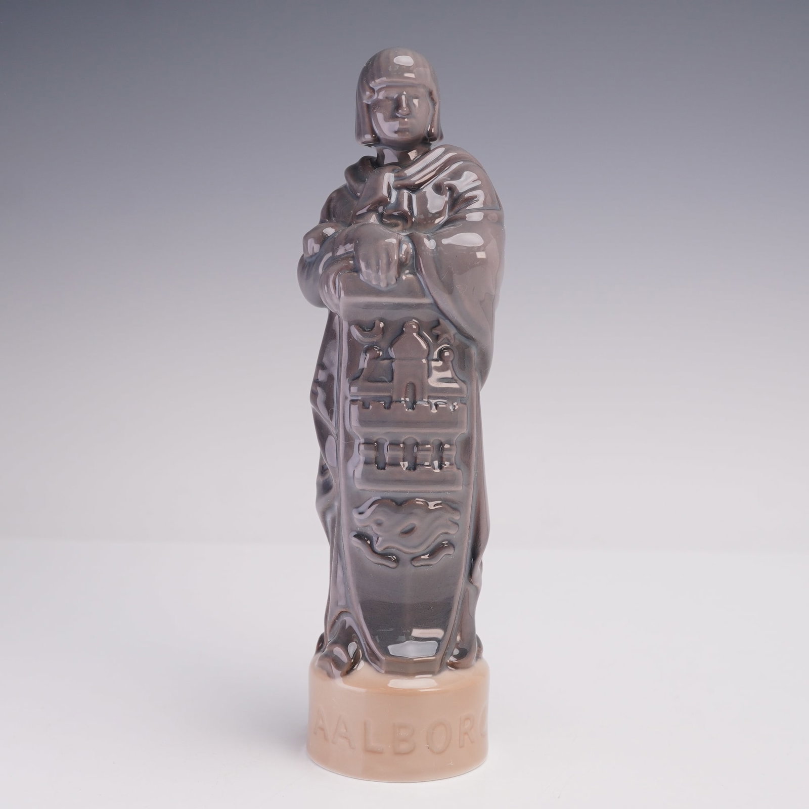 Bing & Grondahl Figurine, Aalborg 2368: A glazed porcelain figurine, the sculptural form depicts a robed figure standing upright with arms folded, detailed with architectural and symbolic reliefs on the front, and set on a circular base ins