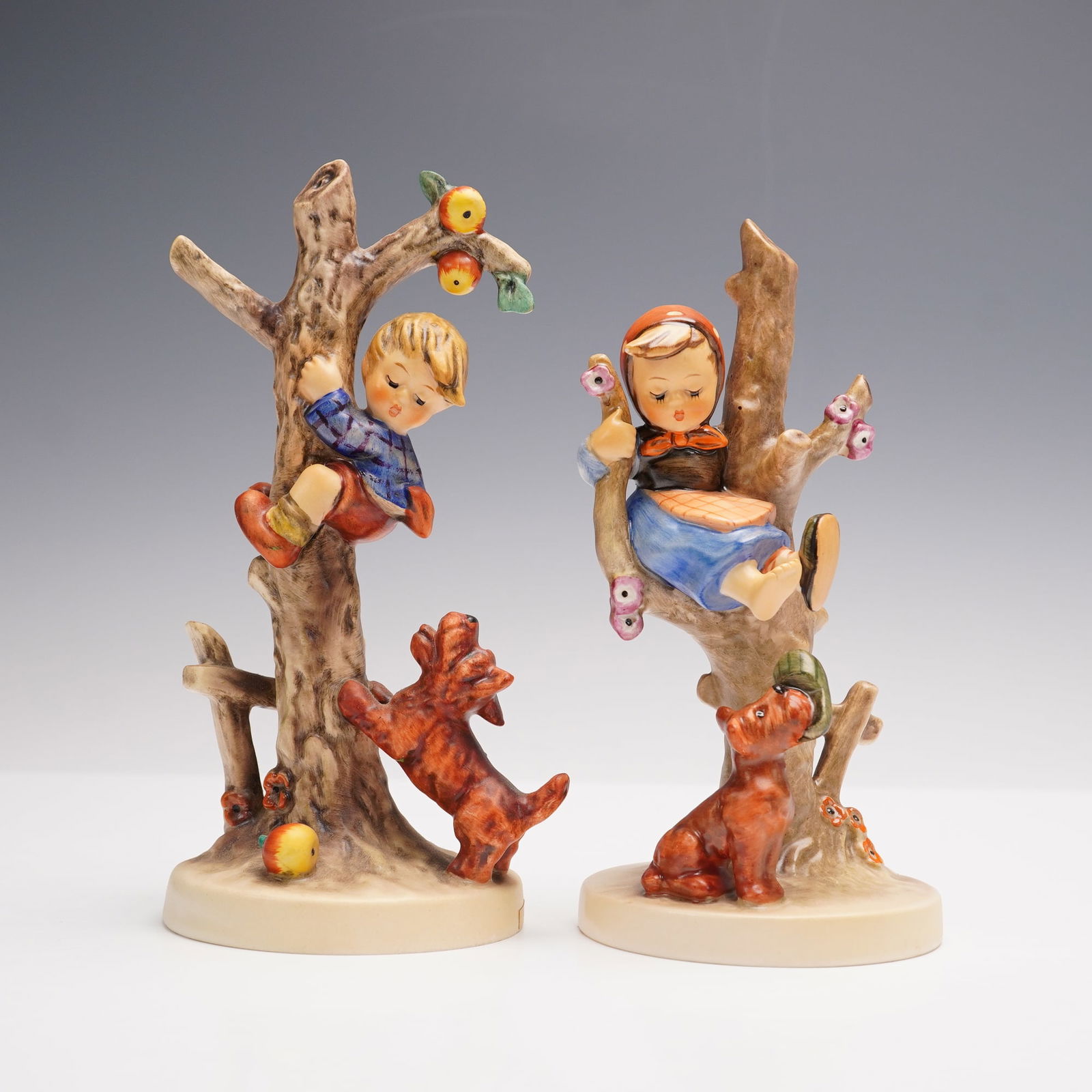 2pc Goebel Hummel Figurines, Children in Trees with Dogs: This pair of Goebel Hummel porcelain figurines features a boy climbing an apple tree while a dog looks up from below, and a girl resting in a flowering tree with a playful dog at the base. Both figure