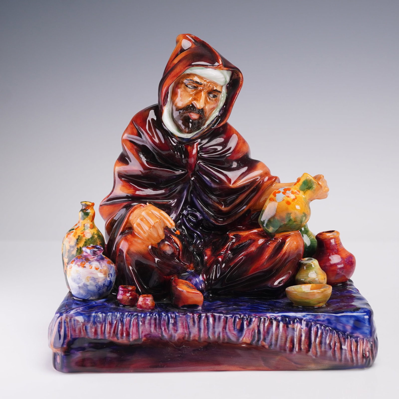 Royal Doulton Figurine, The Potter HN1094: A Royal Doulton figurine depicting a craftsman in a flowing maroon robe, seated among colorful vessels. He holds a vase in one hand and a paintbrush in the other, with rich glossy glazes in deep reds,