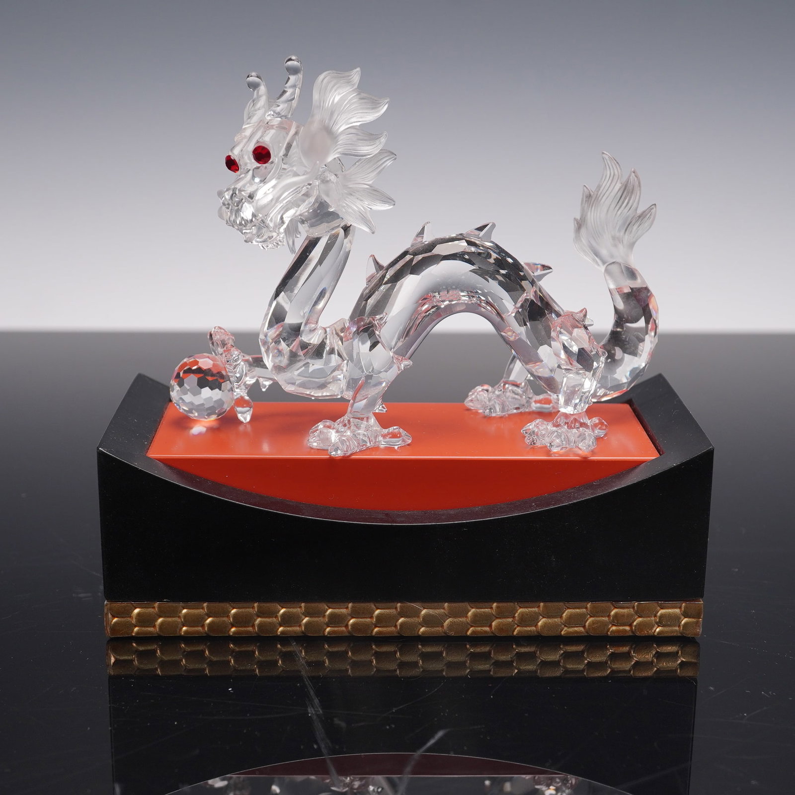Swarovski Annual Edition Fabulous Creatures Crystal Figurine, Dragon: A clear crystal dragon figurine featuring finely detailed facial features, a faceted crystal ball held in one claw, and bright red crystal eyes. The dragon is mounted on a lacquered black and red disp