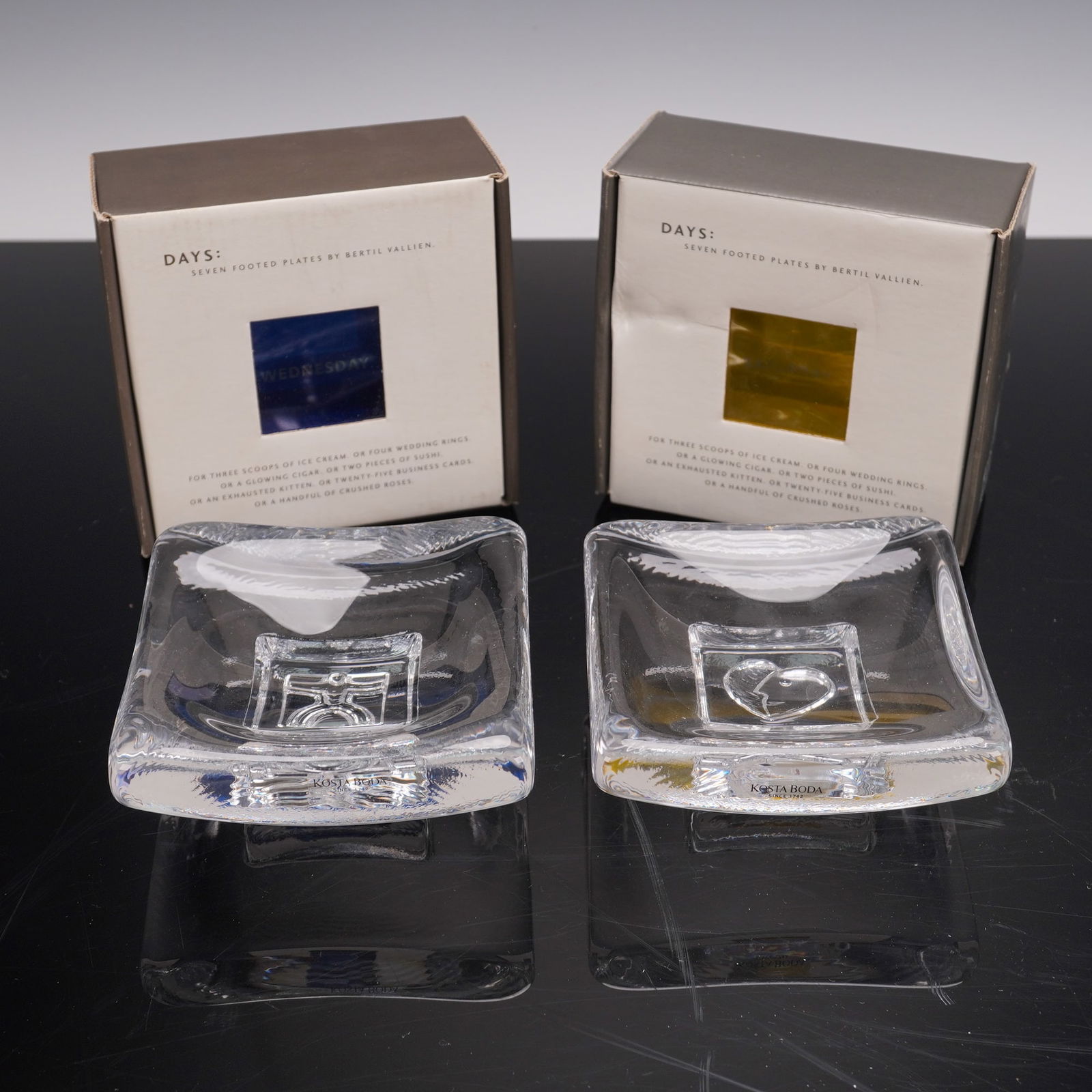 2pc Kosta Boda Days Series Footed Dishes: Pair of Kosta Boda clear art glass footed dishes from the Days series by Bertil Vallien, made in Sweden. Each square dish features a distinct sculptural motif in the center, one depicting a stylized f