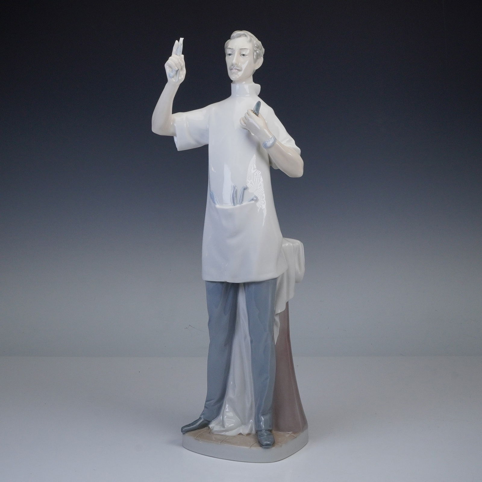 Lladro Porcelain Sculpture, Dentist 1004762: Lladro porcelain sculpture titled Dentist, executed in a glaze finish. Holding a dental instrument with a single tooth in one hand and his folded spectacles in the other, he wears a short-sleeved smoc