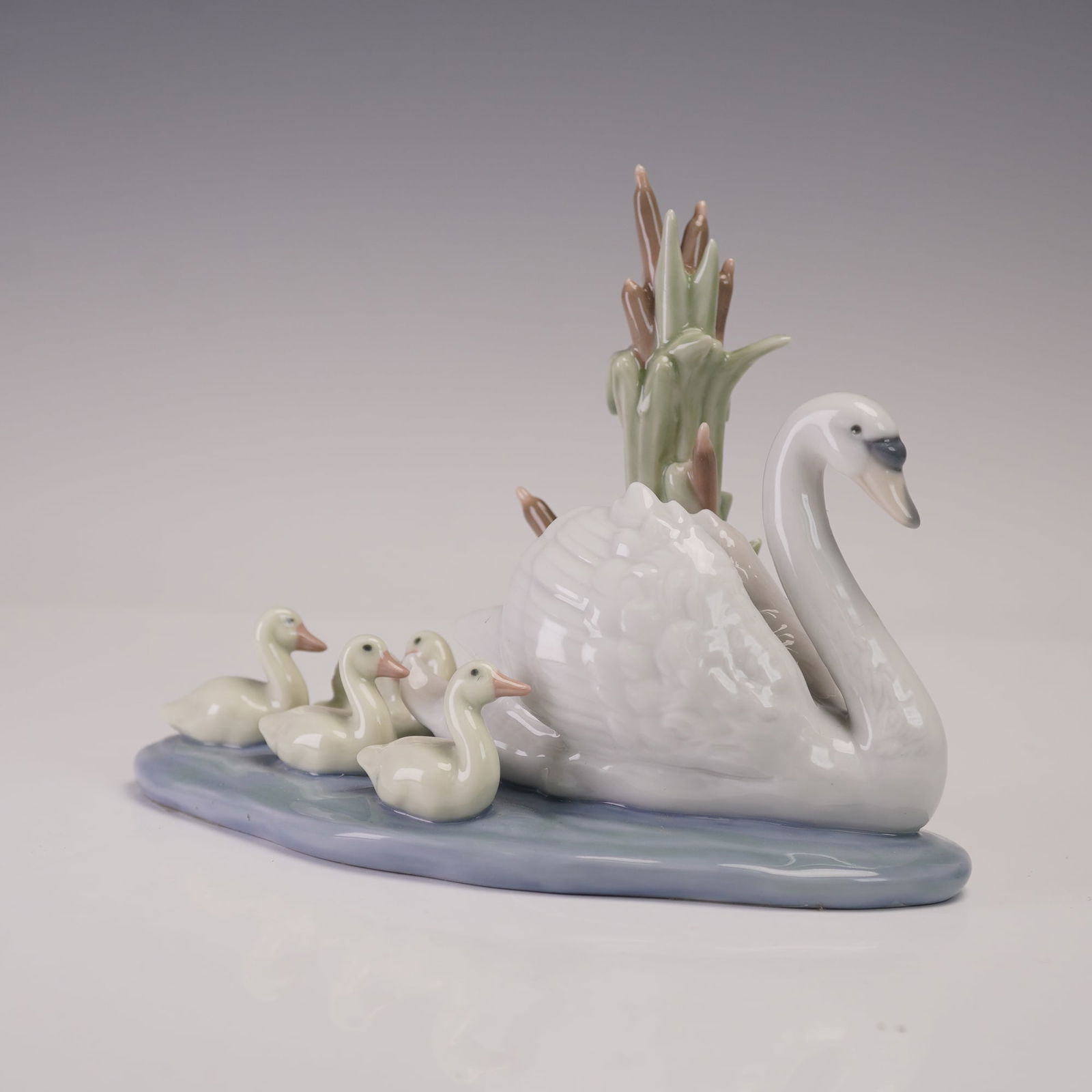 Lladro Porcelain Figurine, Follow Me 1005722: Glossy finish porcelain depicting a serene waterside scene with a swan gliding beside three cygnets, framed by sculpted reeds and cattails rising from the shoreline. Lladro backstamp. This item has it