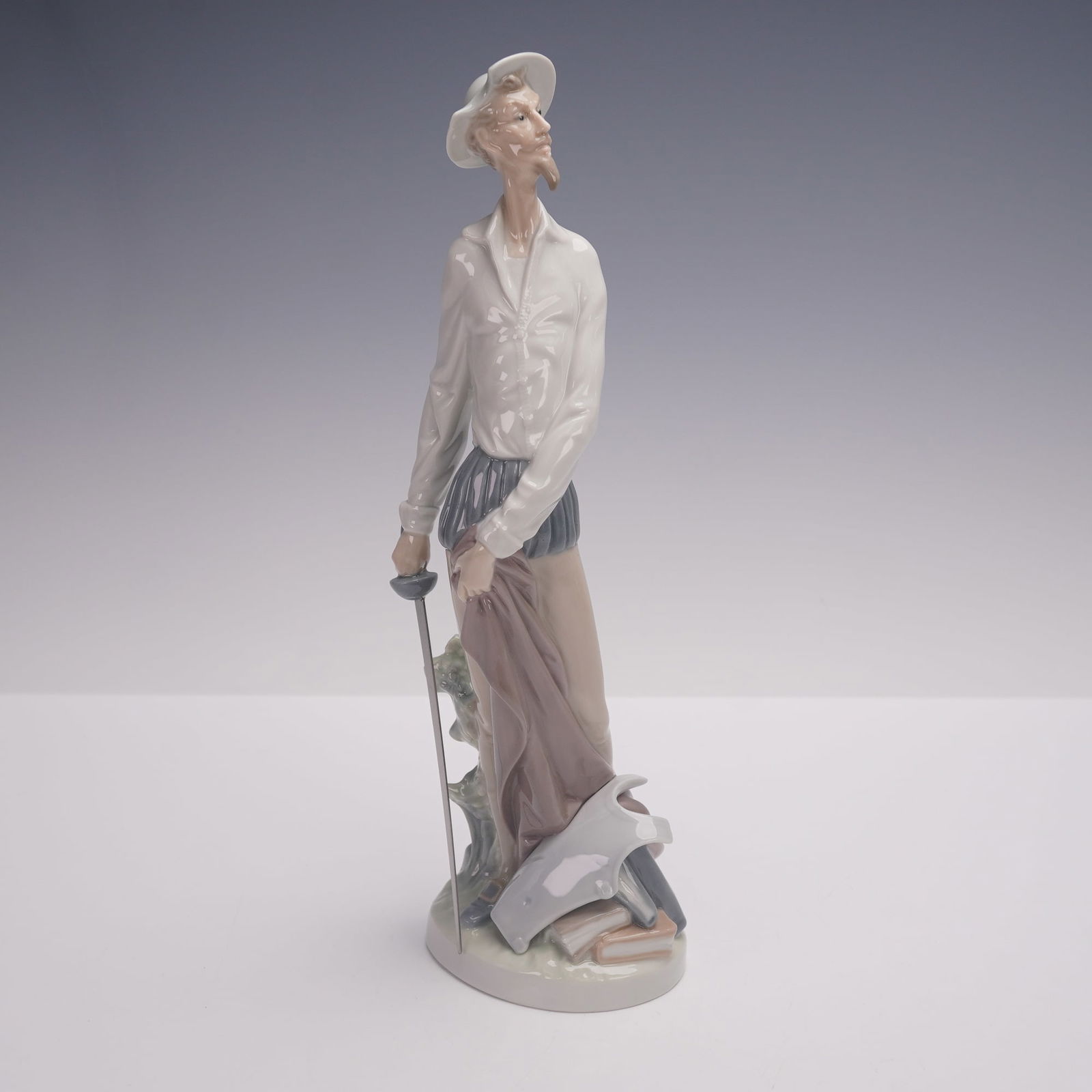 Lladro Porcelain Figurine, Quixote Standing Up 1004854: Lladro porcelain figurine titled Quixote Standing Up, model number 1004854, depicting Don Quixote standing with hands resting on his sword beside a helmet and books. Finished in soft pastel tones with