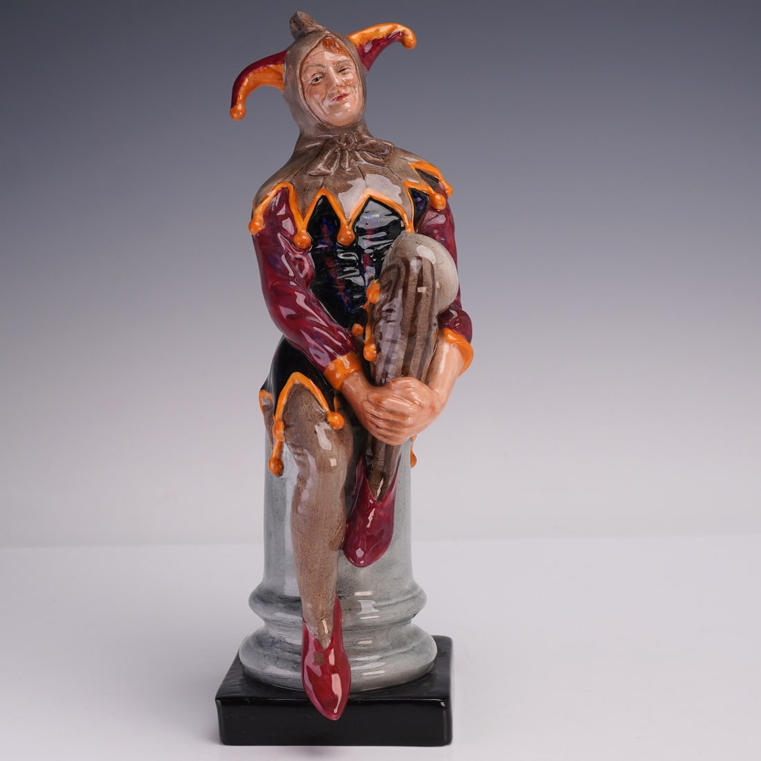 Royal Doulton Figurine, The Jester HN2016: A striking Royal Doulton porcelain figurine titled The Jester HN2016. This piece depicts a colorful court fool seated on a column, dressed in a vibrant costume with maroon sleeves, black bodice, and a