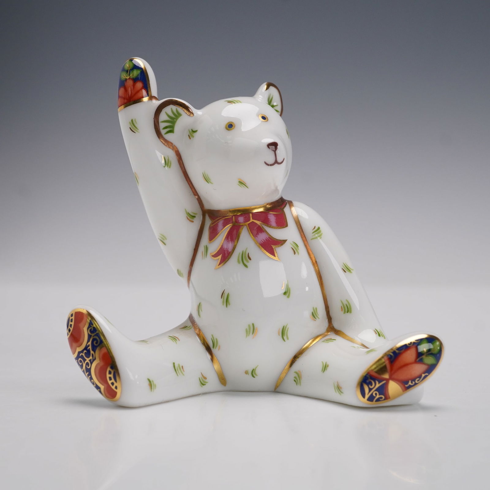 Royal Crown Derby Porcelain Mini Victoria Waving Teddy Figurine: A Royal Crown Derby porcelain miniature figurine depicting a teddy bear named Victoria, designed in a seated position with one arm raised in a wave. The bear is decorated with delicate green strokes a