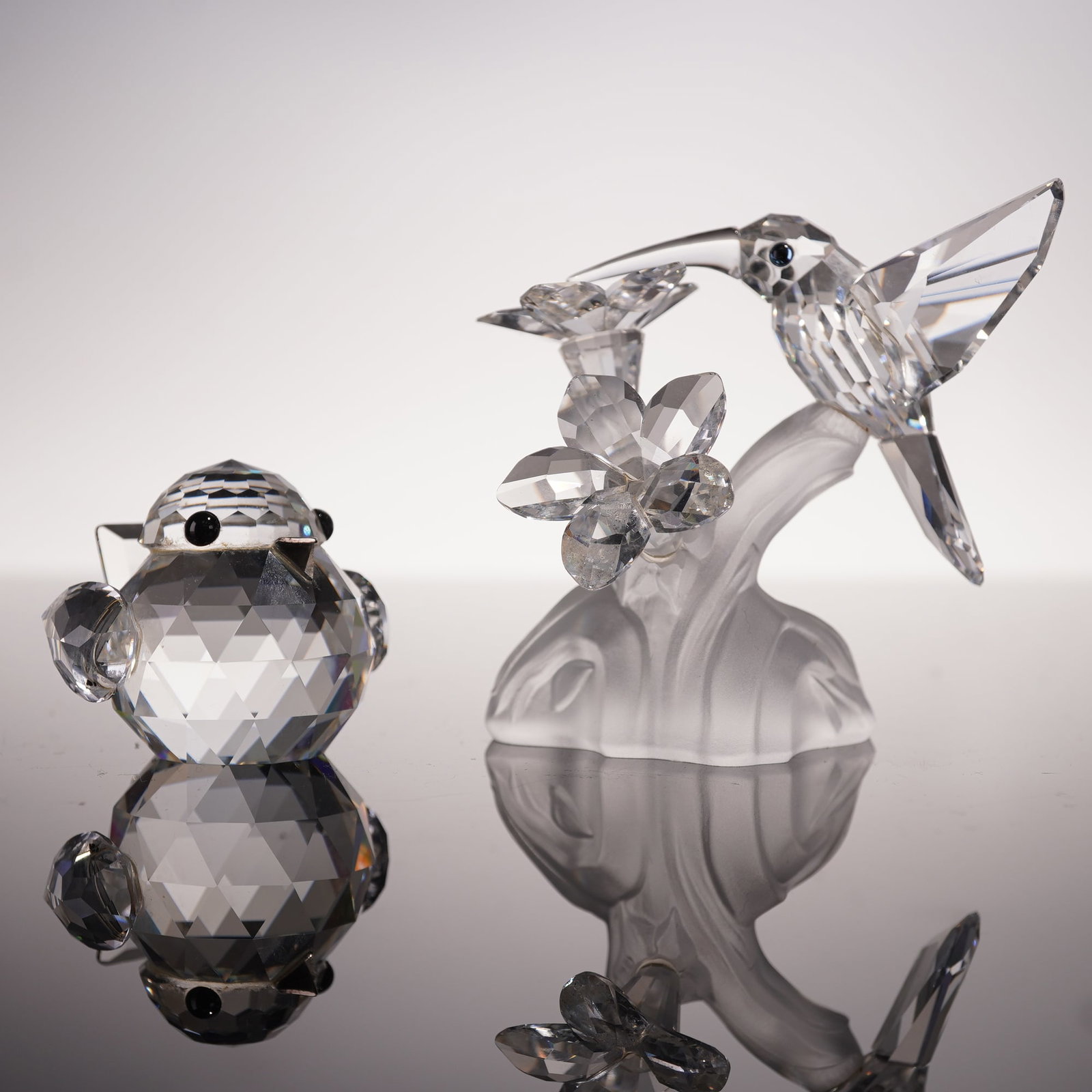 2pc Swarovski Crystal Figurines, Hummingbird & Large Sparrow: Swarovski crystal figurines designed by Claudia Schneiderbauer and Max Schreck. The hummingbird is shown in mid-flight, poised over a faceted crystal flower mounted on a frosted crystal base, highligh