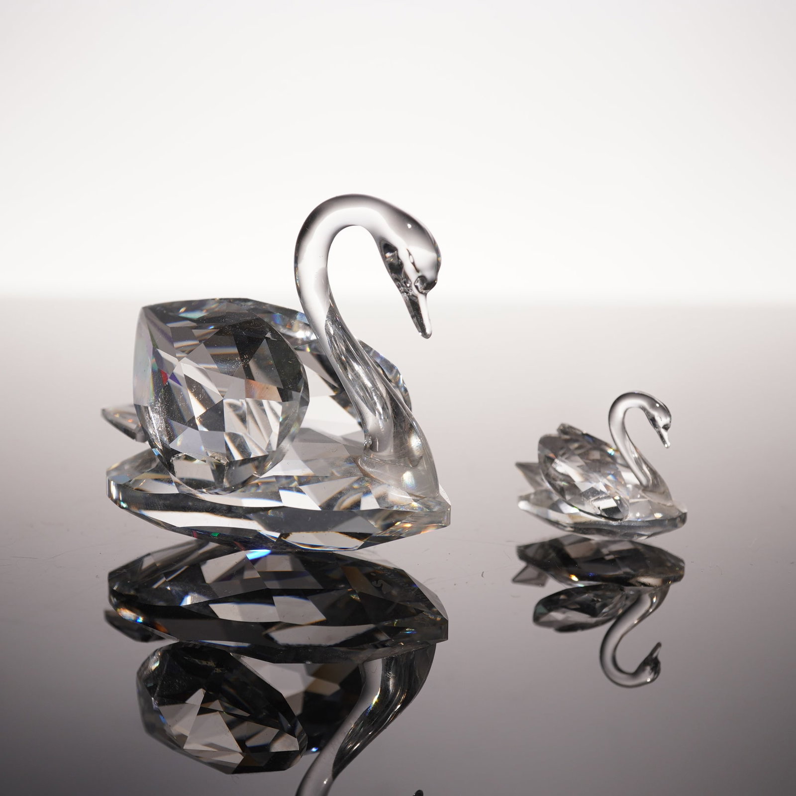 2pc Swarovski Silver Crystal Figurines, Swans: Two silver crystal swan figurines, rendered in faceted crystal, the larger swan displaying broad, multi-angled planes that refract light into prismatic brilliance, while the smaller counterpart mirror