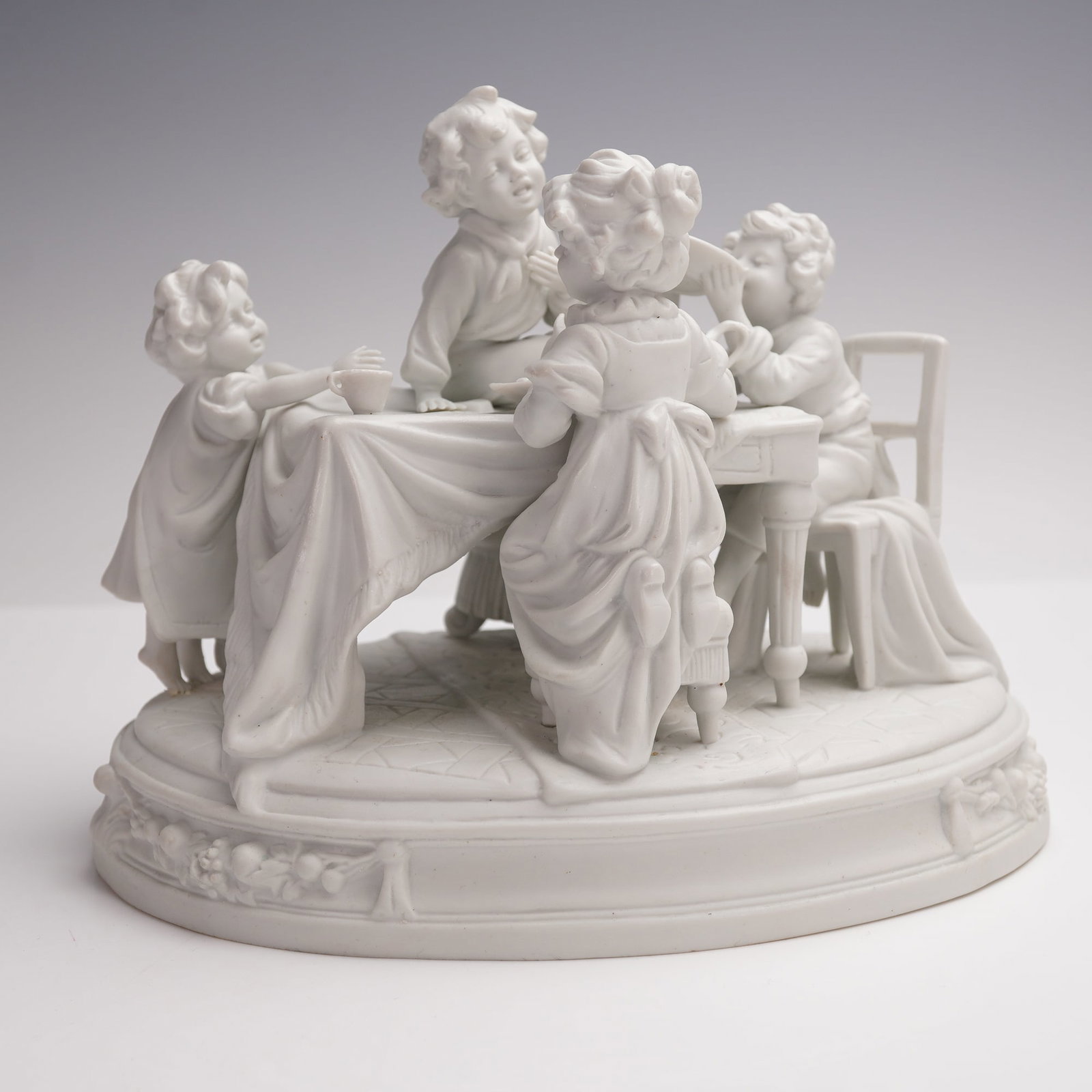 Scheibe-Alsbach Bisque Porcelain Figurine, Children at a Dinner Table: The finely detailed group shows the youngsters engaged in lively activity, with one child perched on a chair, another reaching across the table, and others enjoying food and drink. The scene rests on 