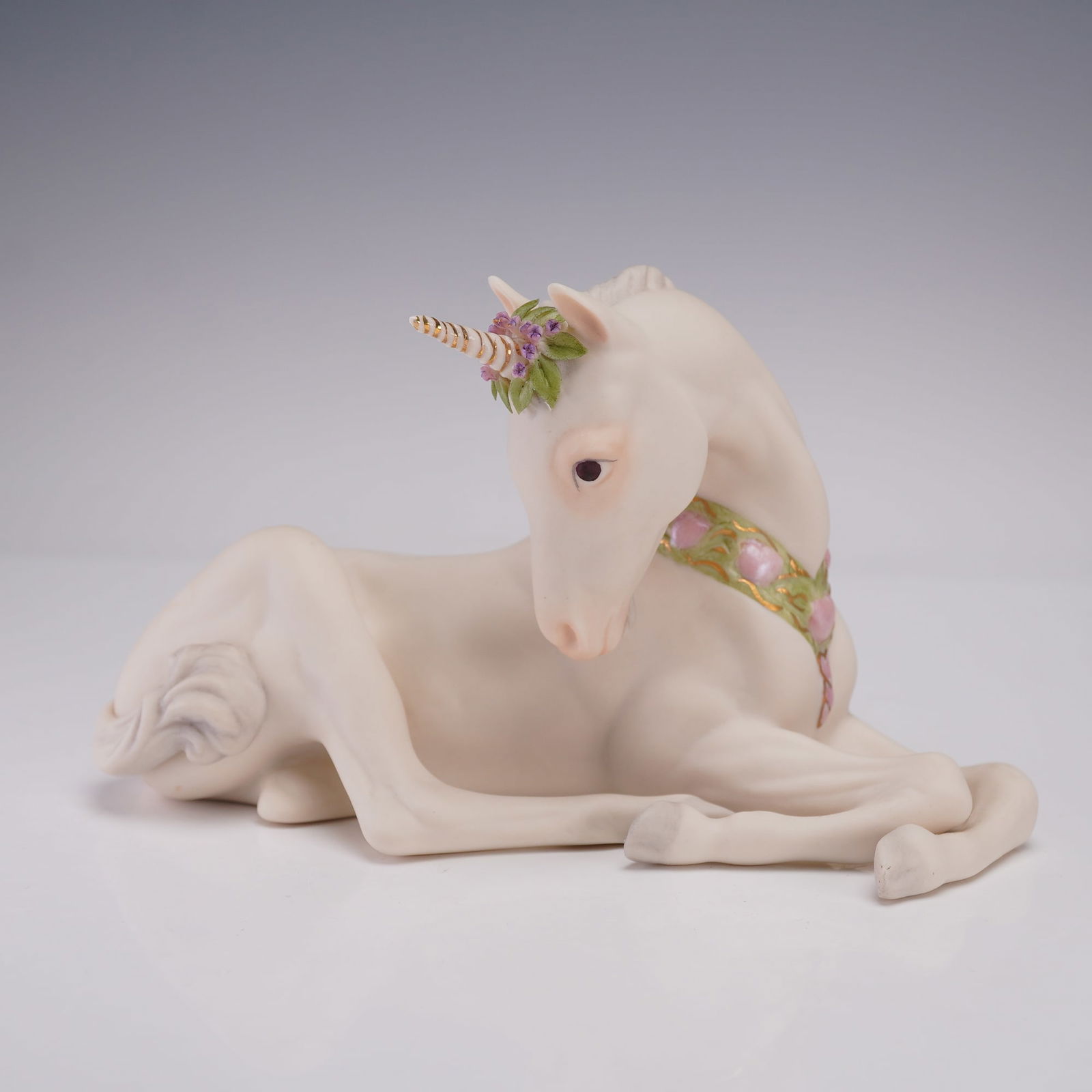 Cybis Porcelain Unicorn Figurine: A Cybis porcelain unicorn shown reclining with its legs folded beneath its body. The unicorn has a delicate pastel floral wreath on its head, a spiraled horn with gilded accents, and a decorative coll