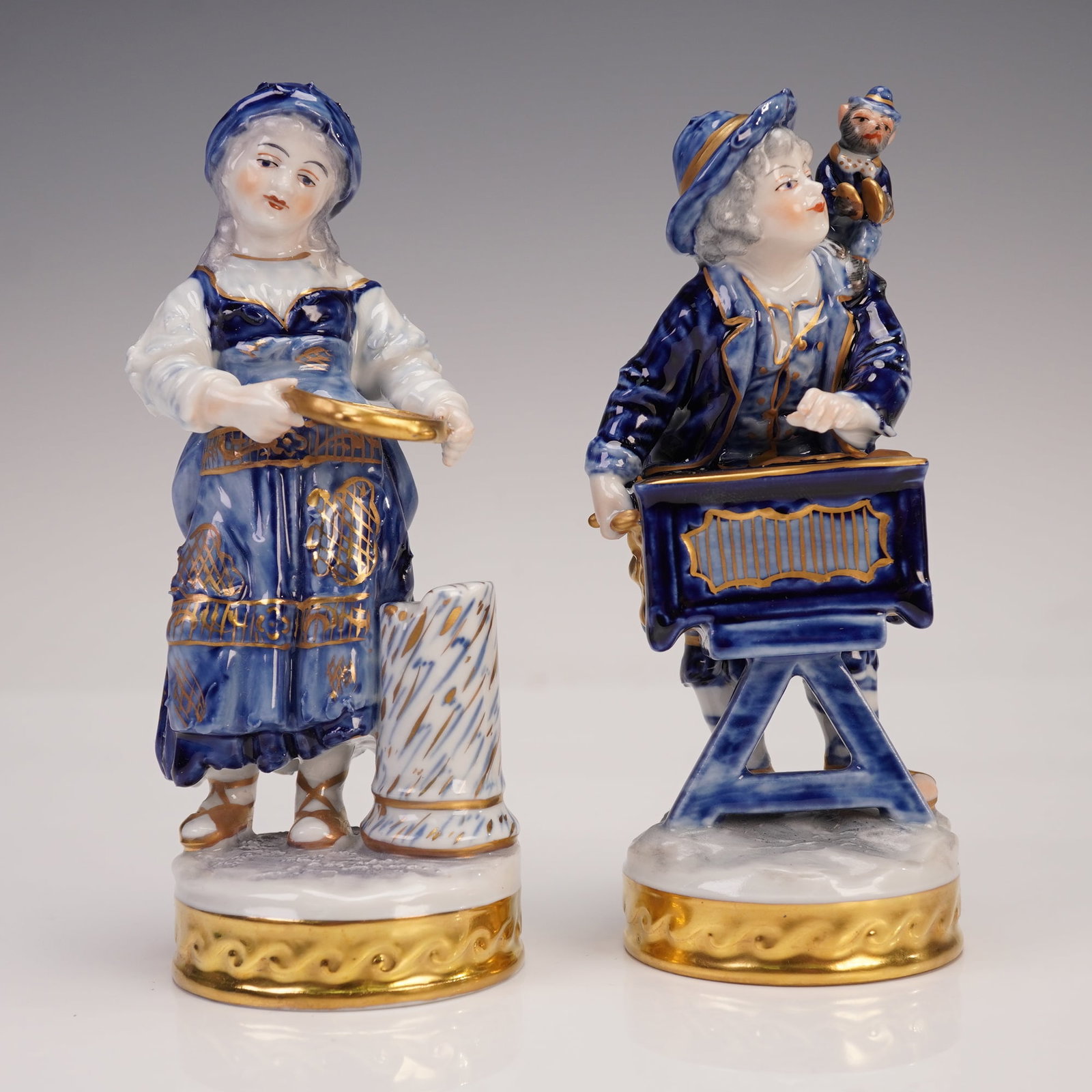 2pc Volkstedt Porcelain Figurines, Boy with Monkey & Waitress: Two hand-painted German porcelain figures attributed to the Aelteste Volkstedter Porzellanmanufaktur, comprising an organ grinder boy with a monkey perched on his shoulder and a waitress with tray bes
