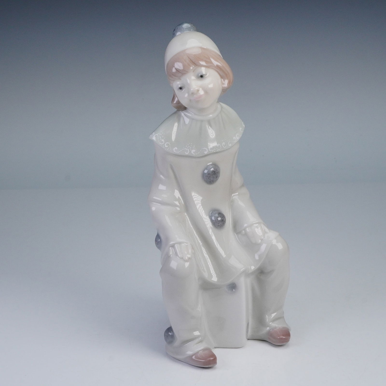 Lladro Figurine, Girl with Dice 1001176: A Lladro porcelain figurine titled Girl with Dice 1001176, depicting a seated clown girl with a wistful expression, dressed in a ruffled collar and costume with large buttons, finished in soft pastel