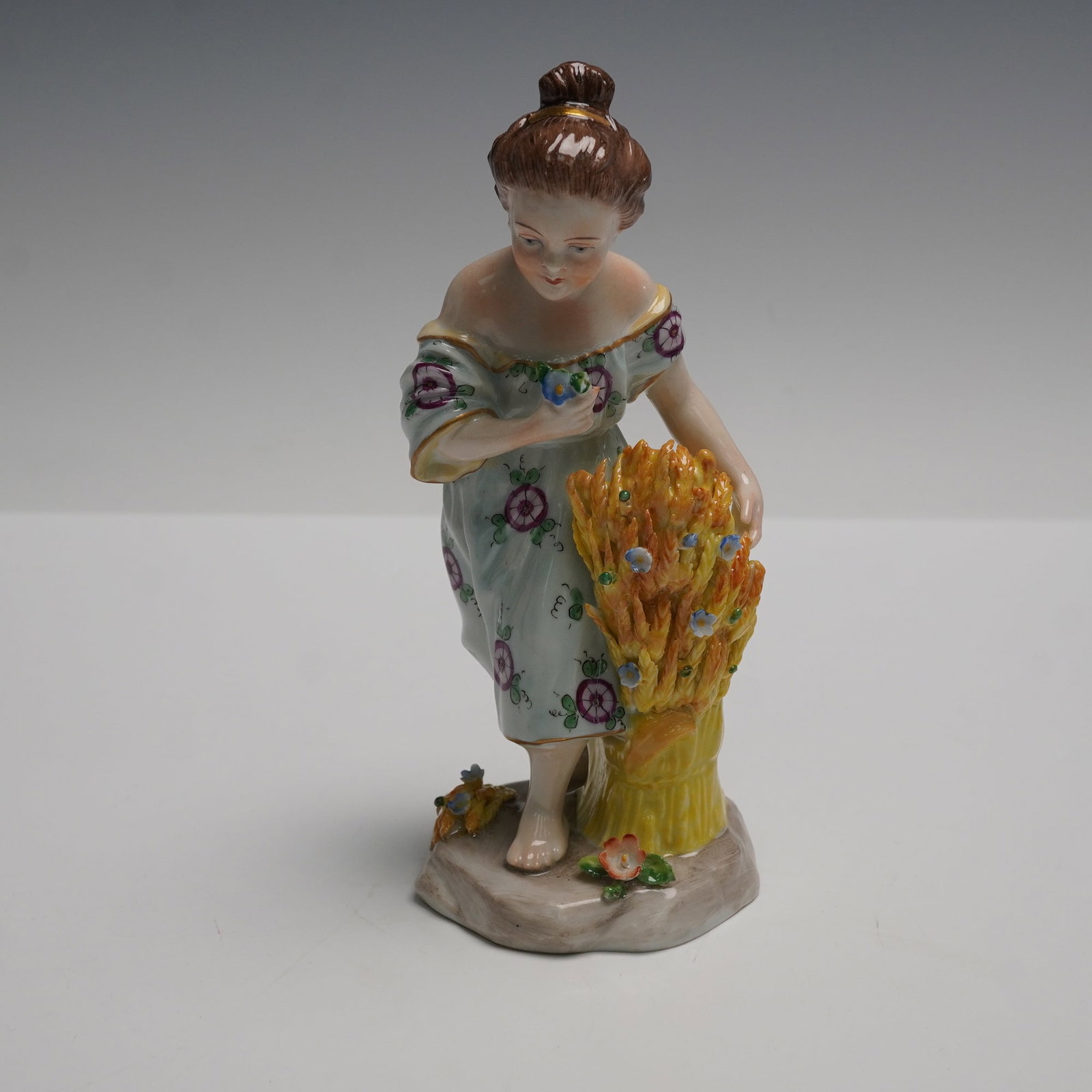 Sitzendorf Figurine, Girl with Wheat: Porcelain figurine depicting a young girl in a floral dress standing beside a bundle of wheat. The figure is delicately hand-painted, showing fine details in the hair, face, and attire, with added tou