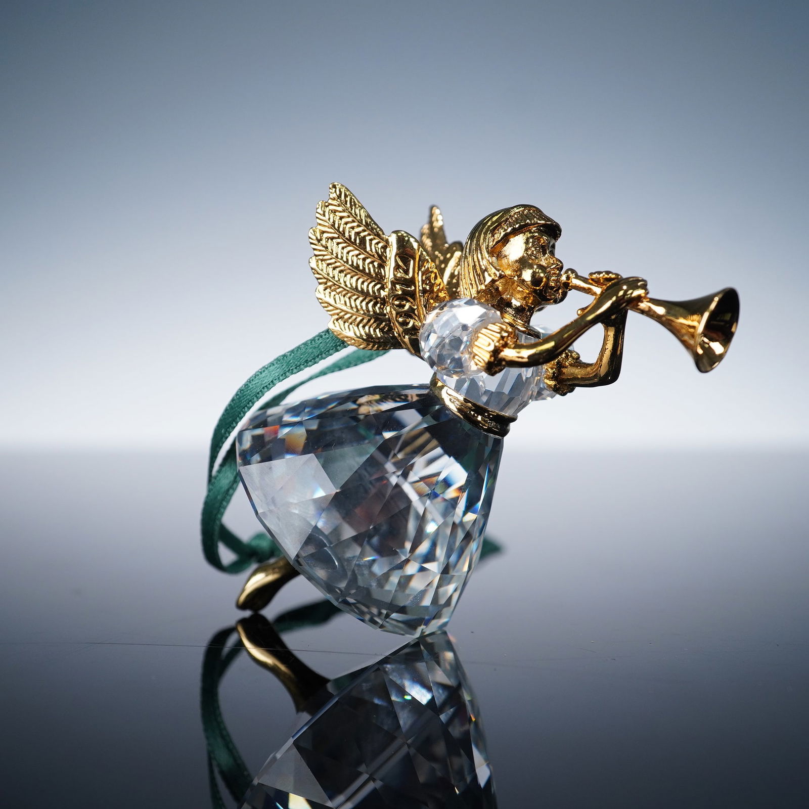 Swarovski Christmas Memories Crystal Ornament, 1997 Angel: A finely crafted ornament featuring a faceted clear crystal body with a gold-tone angel playing a trumpet. The angel's detailed wings and musical instrument are rendered in polished gilt metal, comple