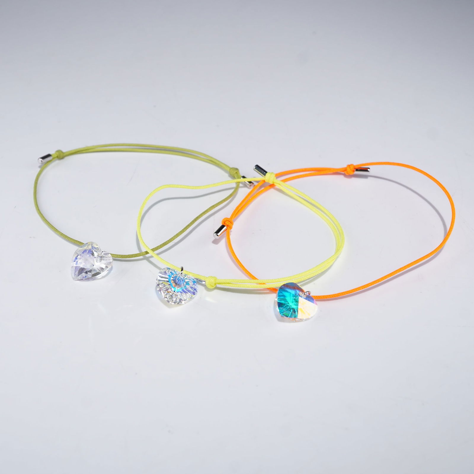 3pc Swarovski Crystal Heart Charm Bracelets: A colorful set of three Swarovski crystal bracelets with faceted heart-shaped crystal charms. The charms display brilliant sparkle and light refraction, paired with adjustable cord bands in green, yel