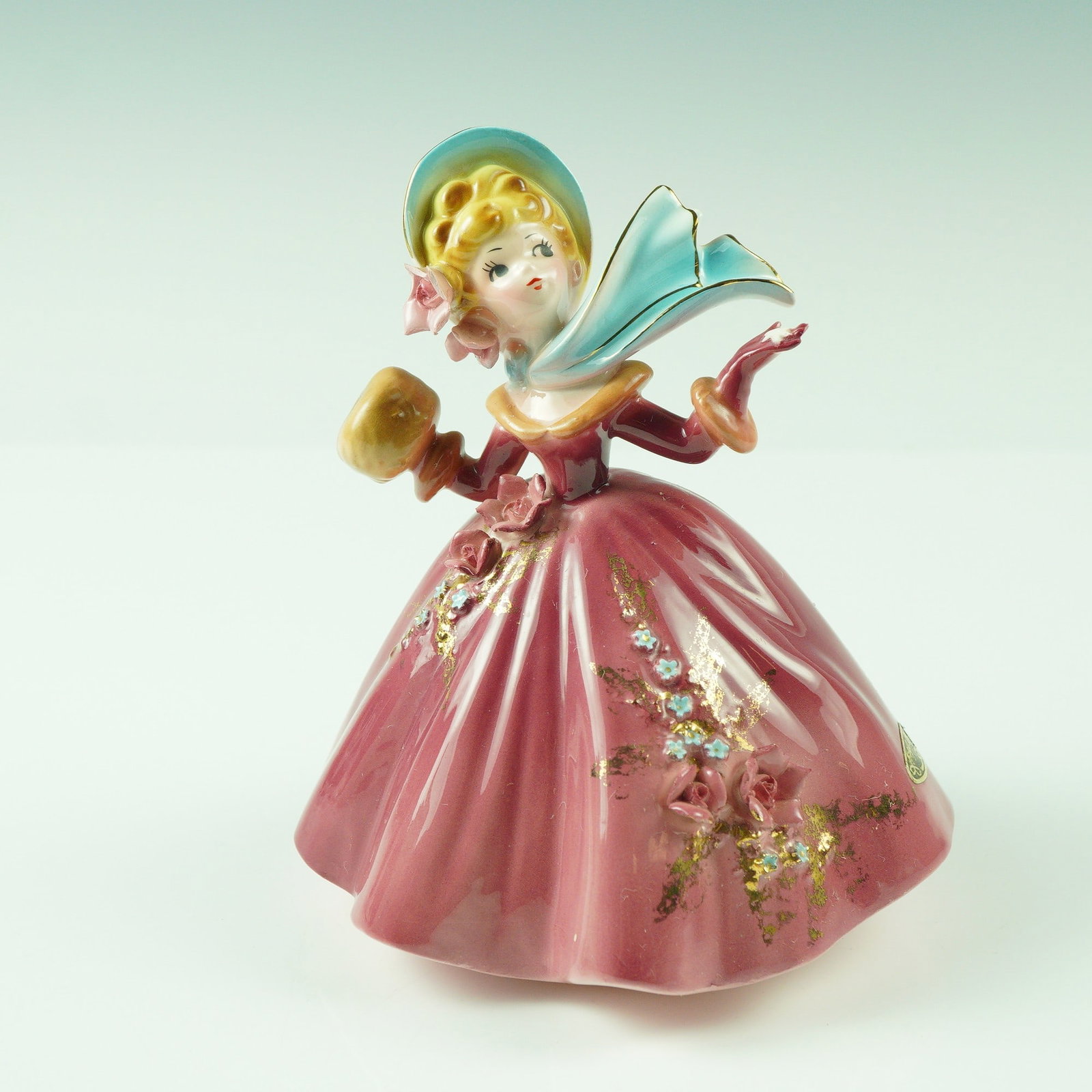 Josef Originals Porcelain Figurine, Winter Girl: This charming Josef Originals figurine depicts a lovely young lady in a flowing rose-colored gown accented with delicate floral details and hand-applied gold highlights. She wears a matching bonnet an