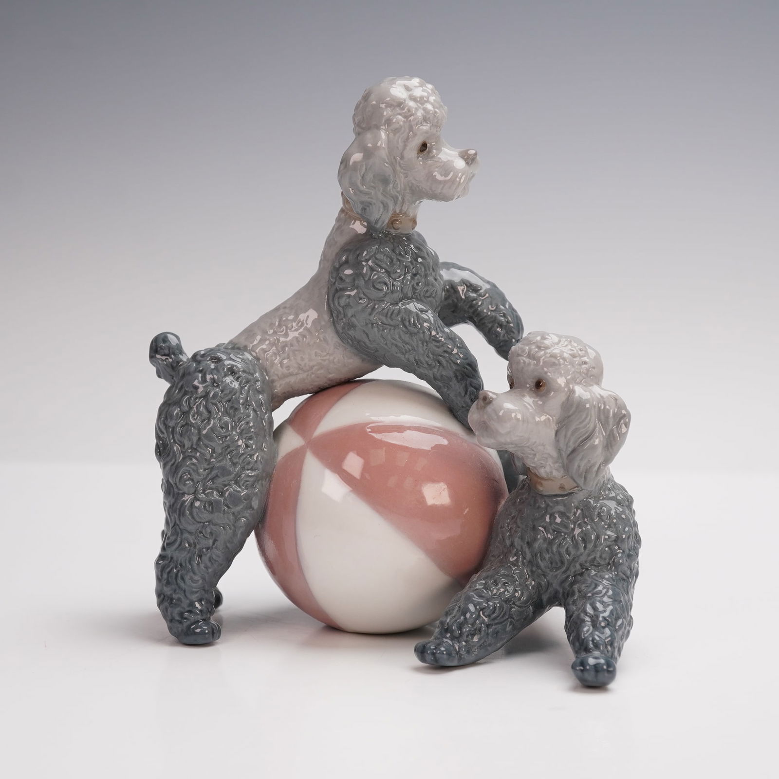Lladro Porcelain Figurine, Playing Poodles, 01001258: A charming Lladro porcelain figurine titled Playing Poodles depicting two finely detailed poodles at play with a large pastel pink and white ball. The pair is modeled in Lladro's signature glossy fini