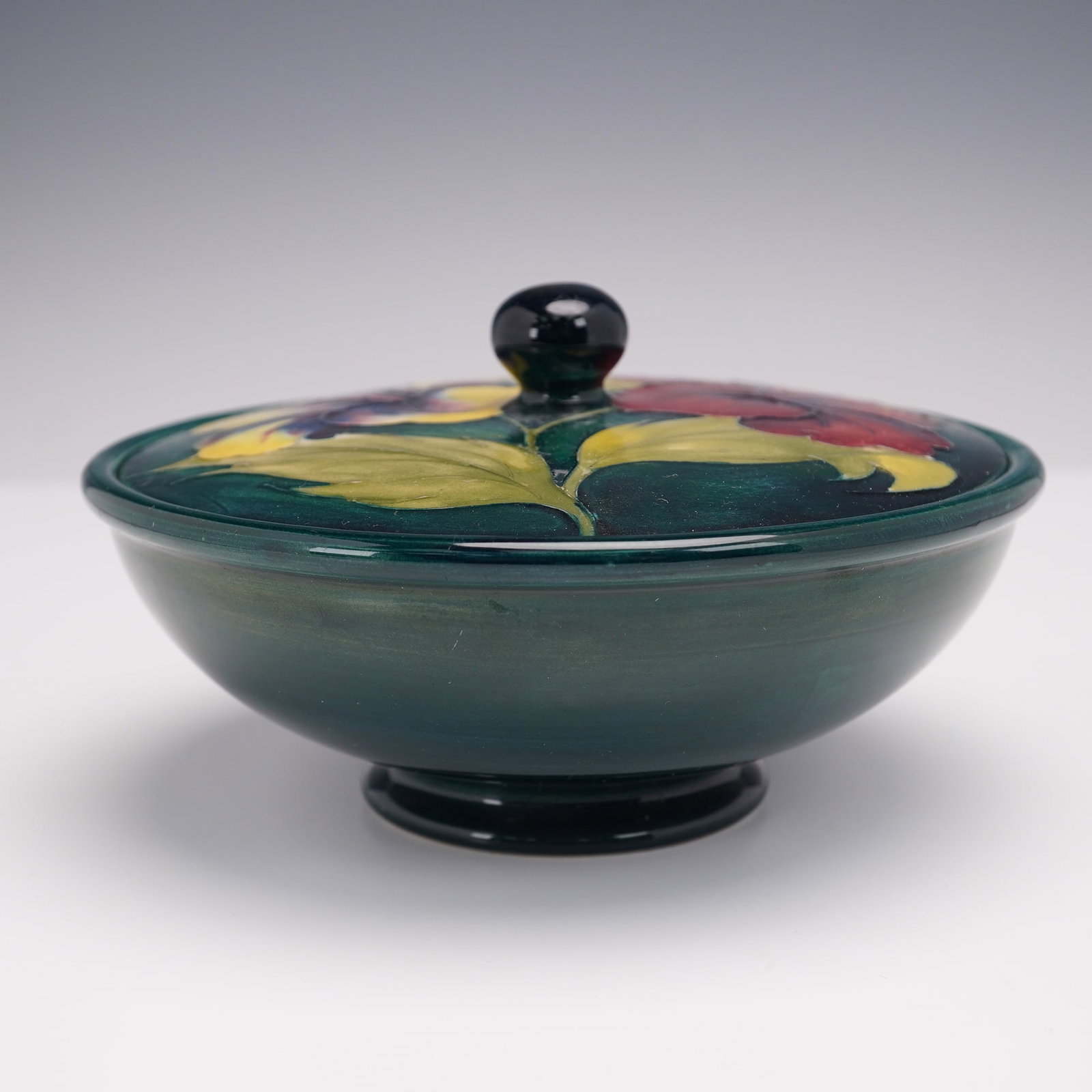 Moorcroft Pottery Covered Bowl, Hibiscus: A vibrant bowl with lid of rounded form raised on a short circular foot. The surface is decorated in the tube lined technique with bold floral motifs in rich shades of crimson and yellow, accented by 