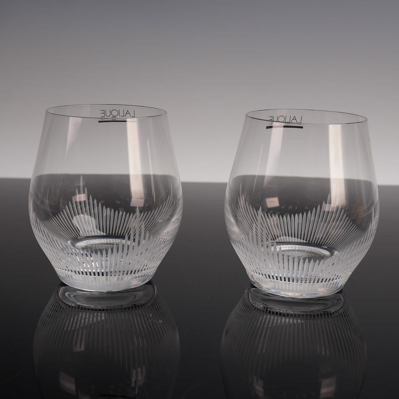 Pair of Lalique Crystal Tumblers, 100 Points: A pair of tumblers from the 100 Points collection. Designed with a sleek rounded form and adorned with finely etched vertical lines radiating upward from the base. Lalique sticker label. Lalique backs