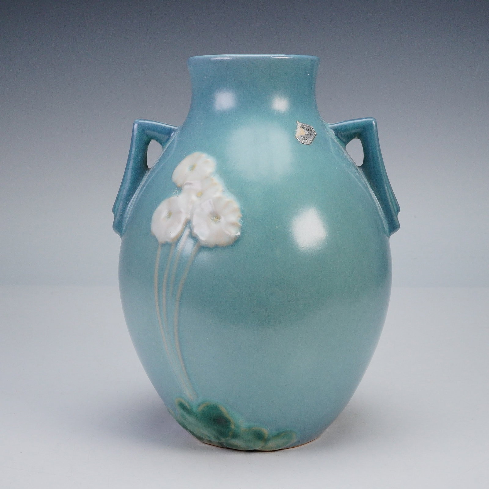Roseville Pottery Blue Vase, Primrose: A vintage Roseville Pottery Primrose vase in a blue glaze with applied white primrose blossoms and green leaf accents. The vase features a rounded body with twin angular handles and subtle embossed de