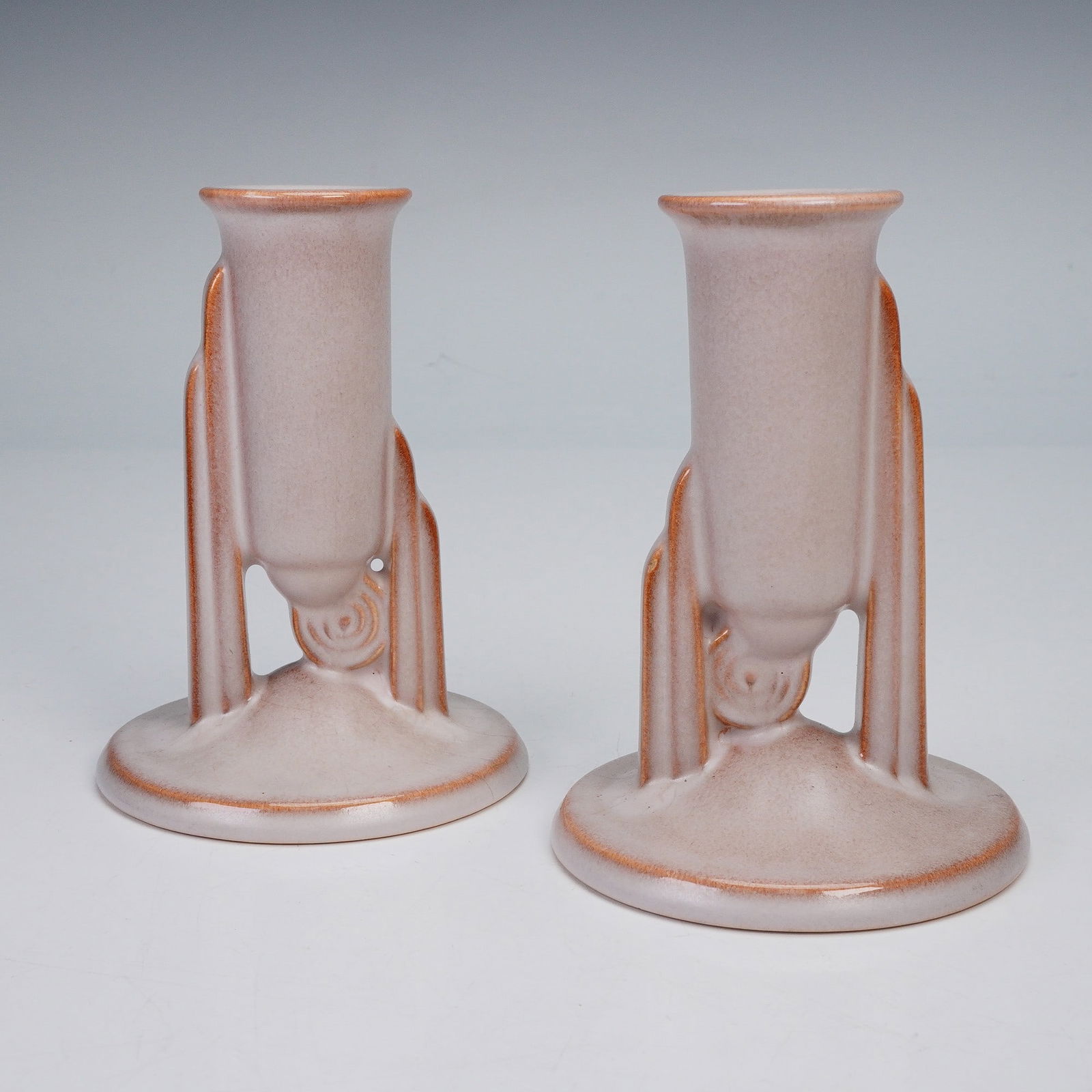 Pair of Roseville Pottery Moderne Art Deco Candle Holders: A set of ceramic candle holders, each featuring a tall cylindrical form with stylized geometric supports rising from a circular base. The soft matte glaze in shades of pale mauve and rose highlights t