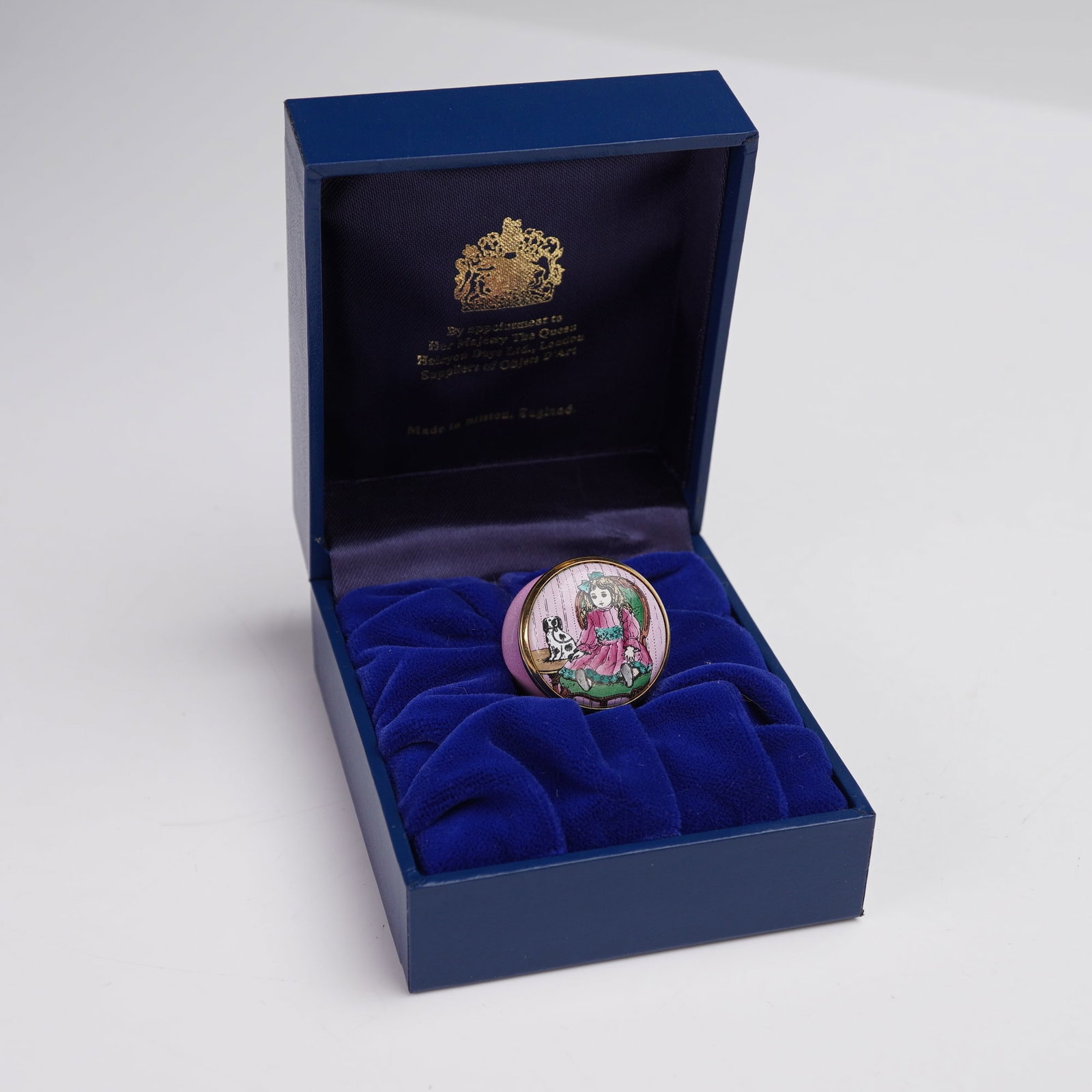 Halcyon Days Enamels Miniature Decorative Box, Victorian Girl Doll and Dog: Enchanting miniature pink enamel decorative box featuring a charming depiction of a doll in a pink dress accompanied by a small black-and-white dog, set against a detailed background. The round enamel