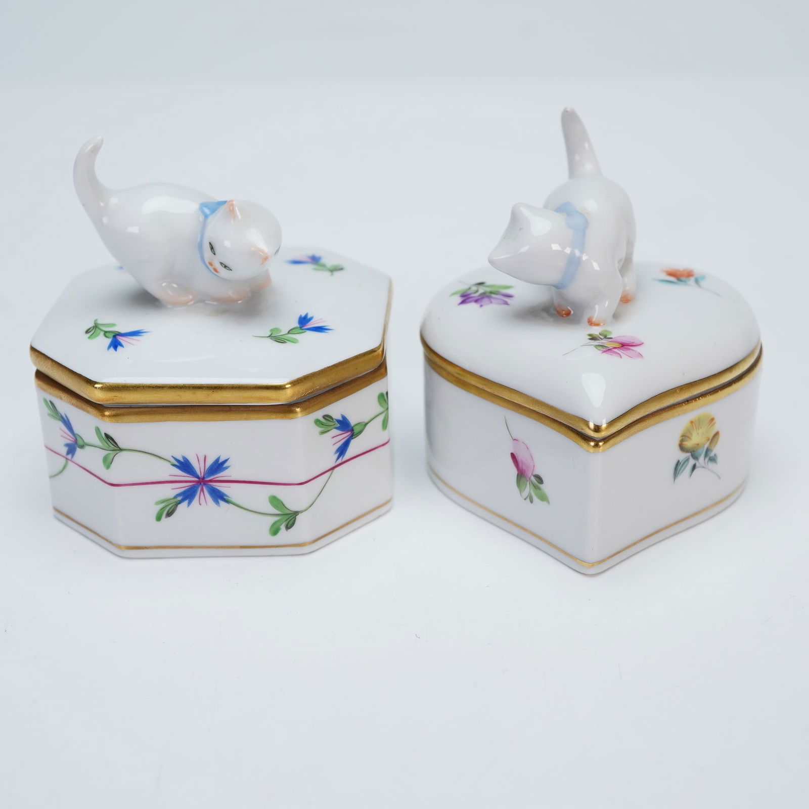 2pc Herend Porcelain Decorative Lidded Boxes: Two adorable porcelain decorative boxes each surmounted by a playful white cat with a blue collar. The hexagonal box is adorned with cornflower motifs, while the heart-shaped box is embellished with d