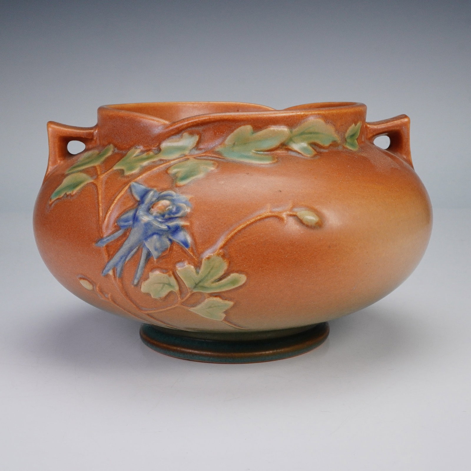 Roseville Pottery Columbine Vase, 655-5: A round art pottery vase, featuring a warm orange ground with raised decoration of green leaves and blue columbine blossoms in soft naturalistic tones. The piece has twin pierced handles at the should