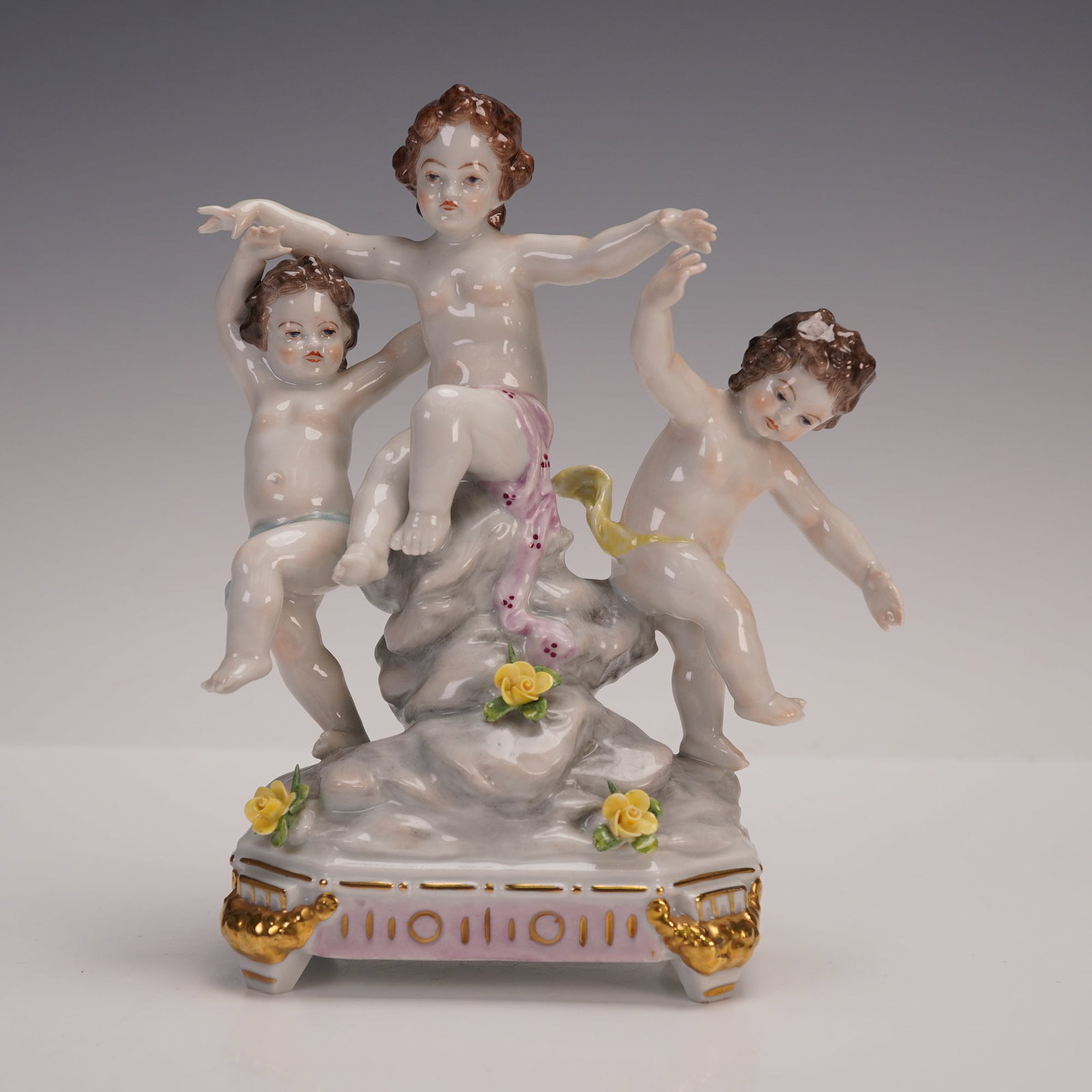 Volkstedt Porcelain Cherubs with Flowers Figurine: This Volkstedt porcelain figurine depicts three playful putti posed around a rocky base accented with applied yellow flowers. The figures are modeled with expressive faces and fine detail, highlighted