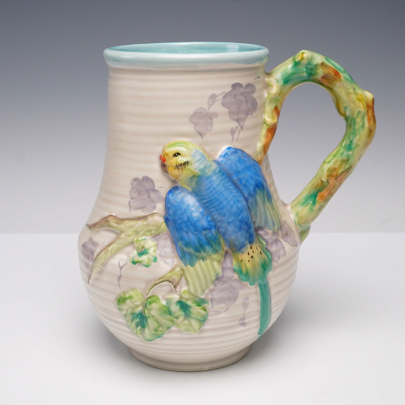Clarice Cliff Newport Pottery Budgerigar Jug 850: A Clarice Cliff Newport Pottery jug featuring a molded and hand-painted design of a brightly colored budgerigar perched on a leafy branch. The handle is formed as a naturalistic branch with multi-colo