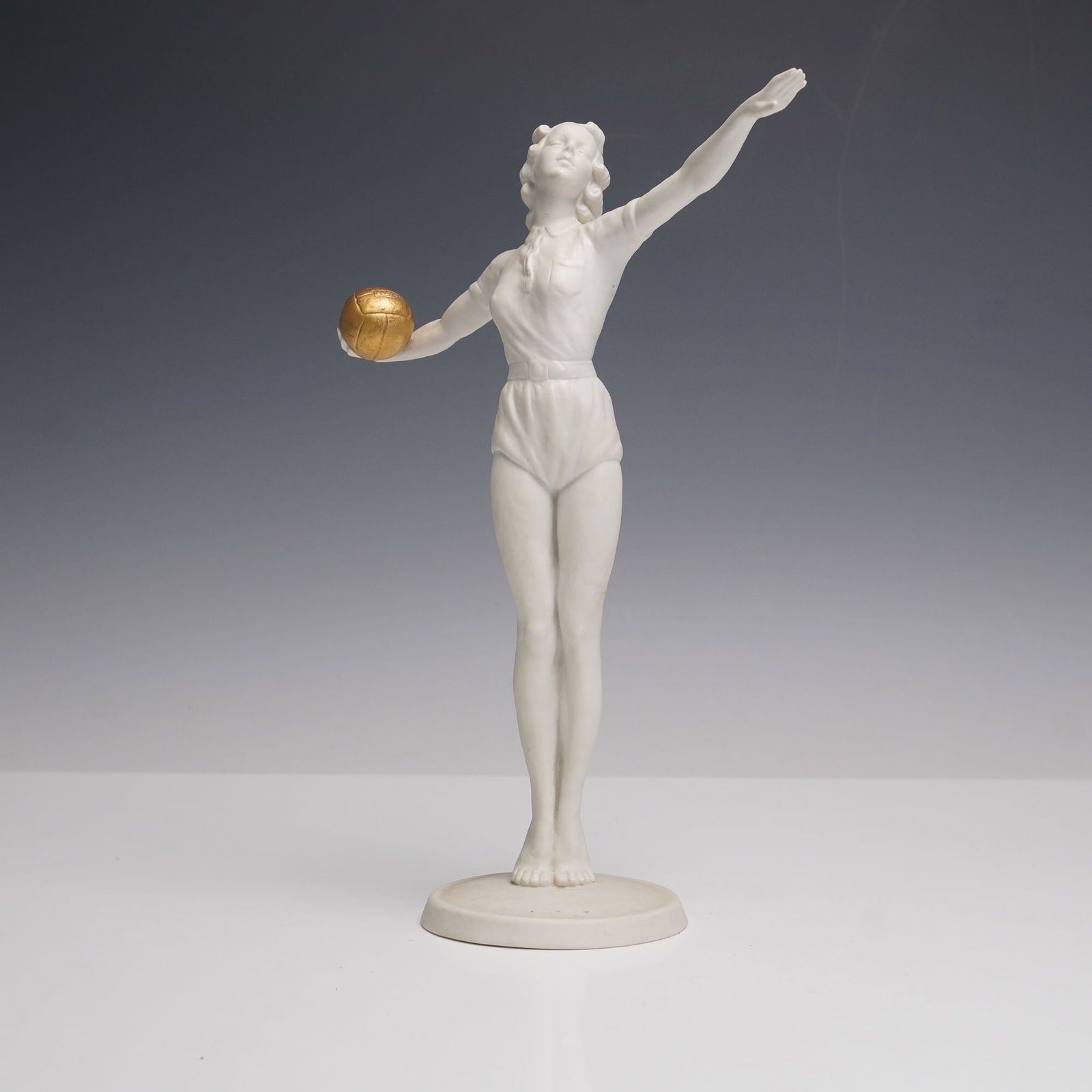 Schaubach Kunst Bisque Figurine, Female Volleyball Player: A finely modeled Schaubach Kunst bisque porcelain figurine depicting a female volleyball player in athletic pose, holding a gilt ball in one hand with the other arm raised. The matte white surface hig