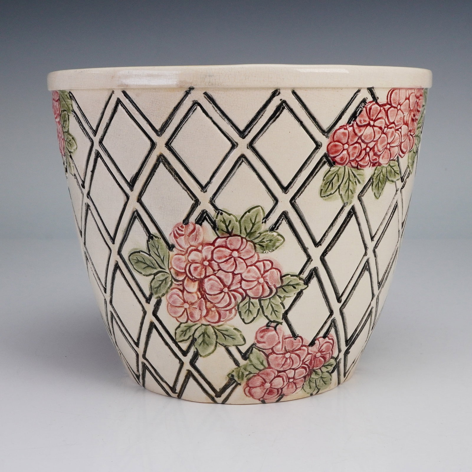 Weller Pottery Jardiniere with Floral Trellis Design: A finely crafted jardiniere from Weller Pottery featuring a raised trellis pattern adorned with clusters of pink blossoms and green leaves. The hand-painted detail and textured surface highlight the b
