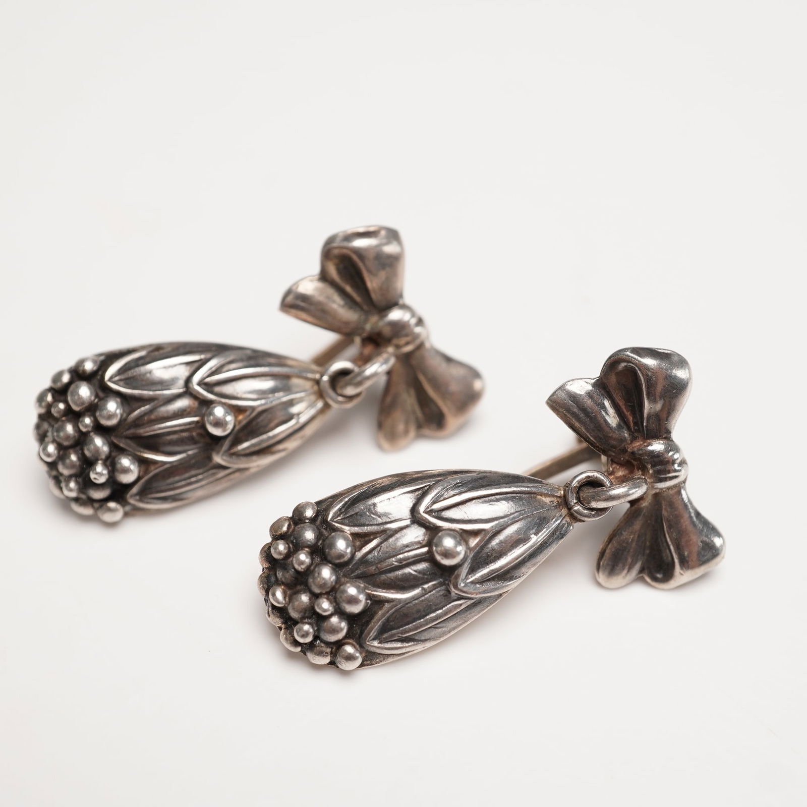 Mexican Sterling Silver Bow with Leaf & Berry Earrings: A pair of Mexican sterling silver earrings crafted with screw-back closures, each designed with an elegant bow surmount suspending a teardrop form decorated with leaf and berry motifs. Stamped STERLIN