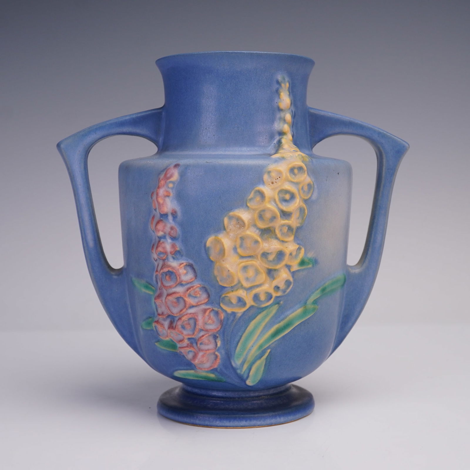 Roseville Pottery Foxglove Blue Vase, 46-7: A double-handled art pottery vase in the Foxglove pattern. The piece features raised floral motifs in shades of yellow and pink against a matte blue background, with green leaves accenting the design.