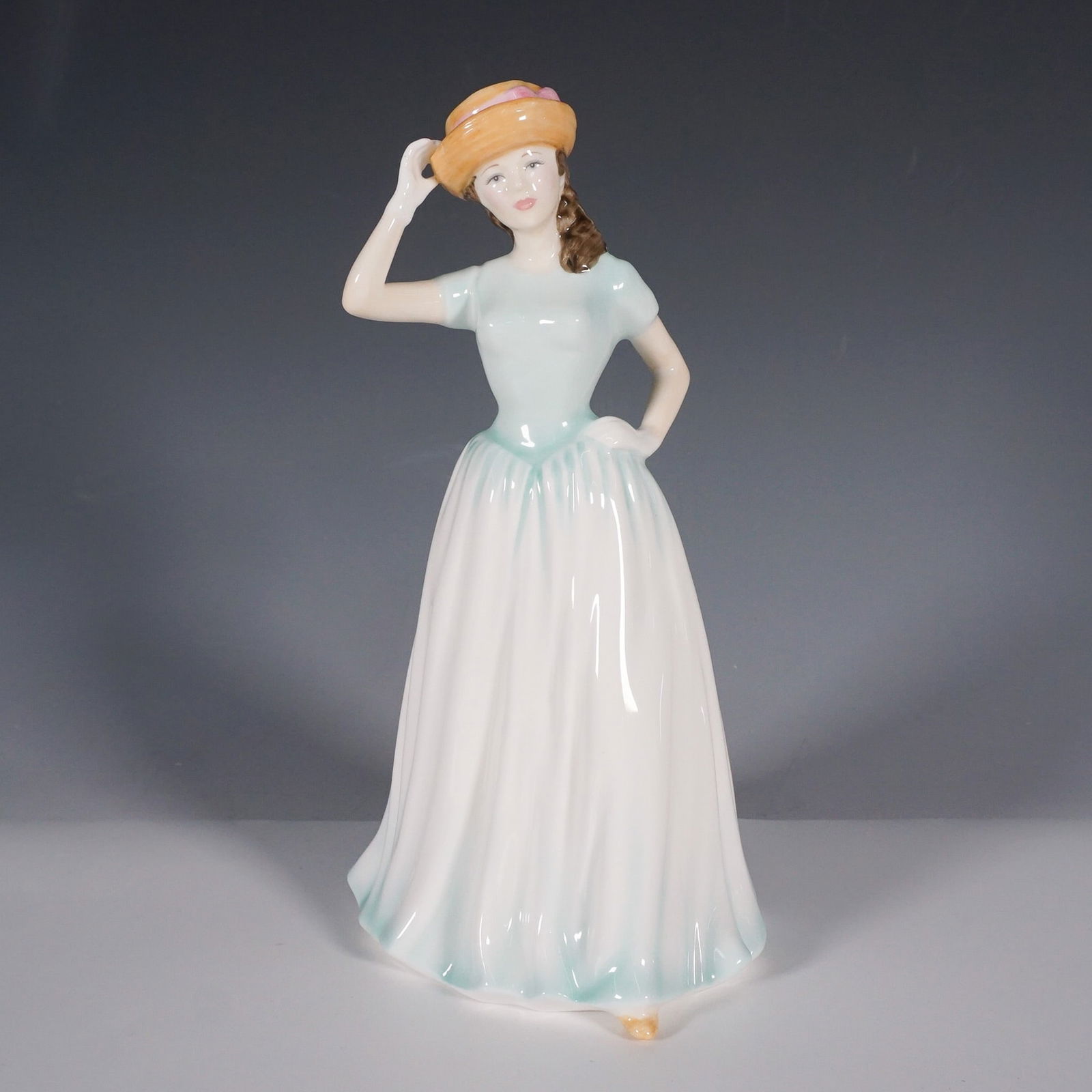 Royal Doulton Figurine Happy Birthday HN4308: This Royal Doulton porcelain figurine, titled Happy Birthday (HN4308), showcases a young woman in a flowing pastel gown with a soft gradient from white to aqua. She wears a charming hat with delicate