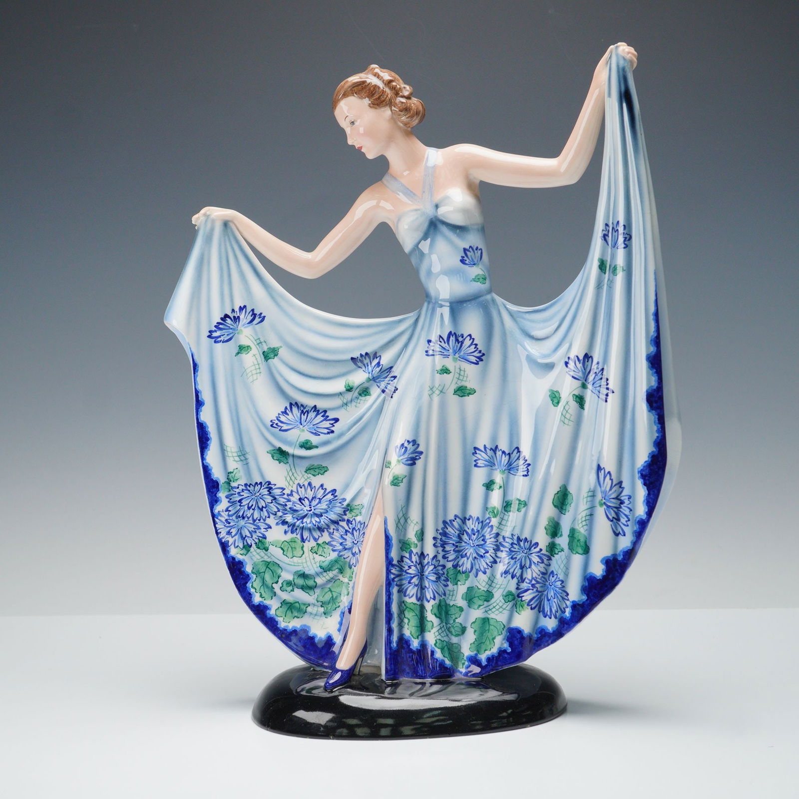 Goldscheider by Stefan Dakon Colorway Figurine: Depicting a young woman posed gracefully while holding out the sides of her long open skirt. The figure wears a strapped bandeau-style bodice and a flowing dress decorated with detailed floral motifs