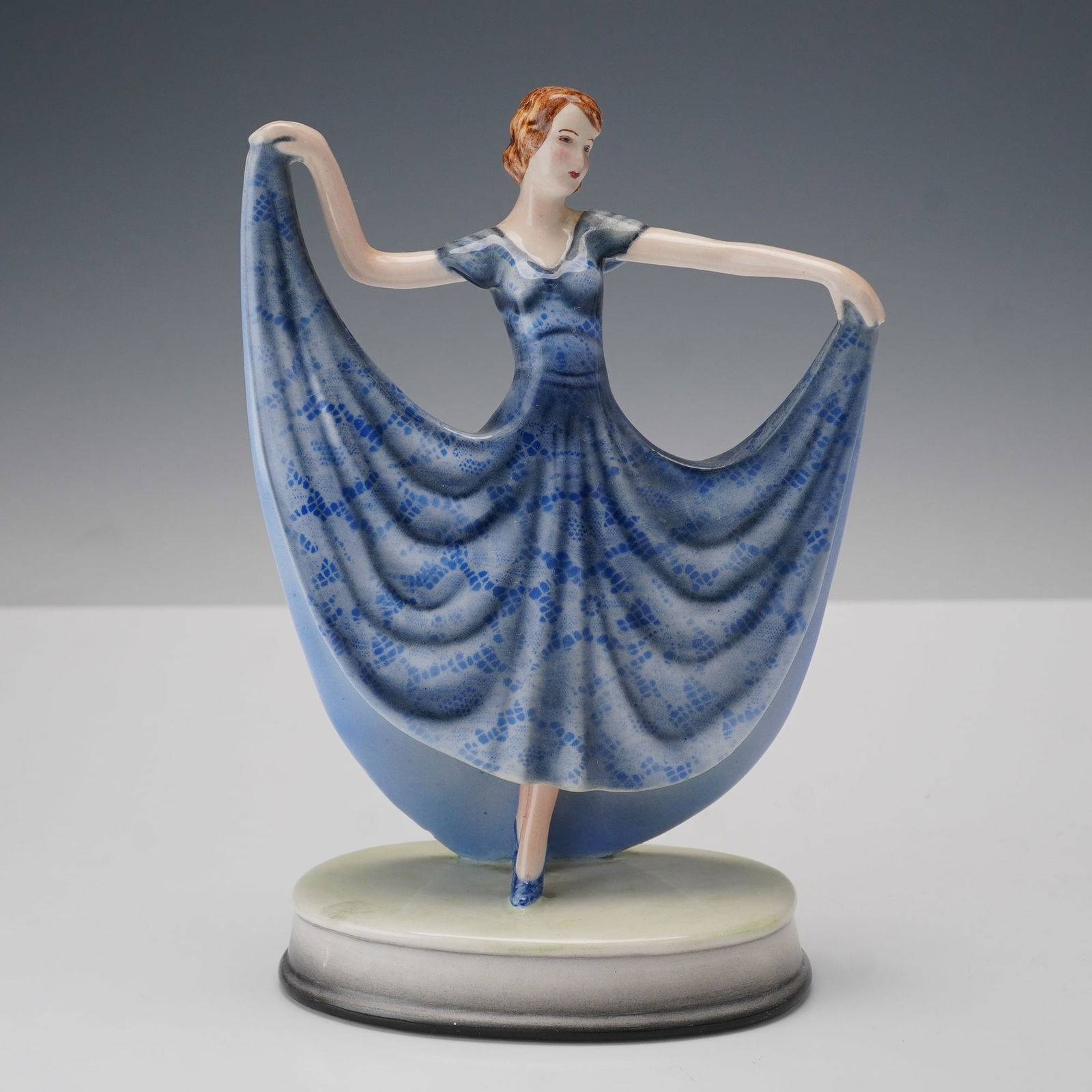 Goldscheider by Stefan Dakon Figurine, Nora: Depicting a woman posed in a flowing blue dress with lace-pattern detailing, her arms outstretched holding the edges of her skirt. The hand-painted porcelain figure stands on an oval base and bears th